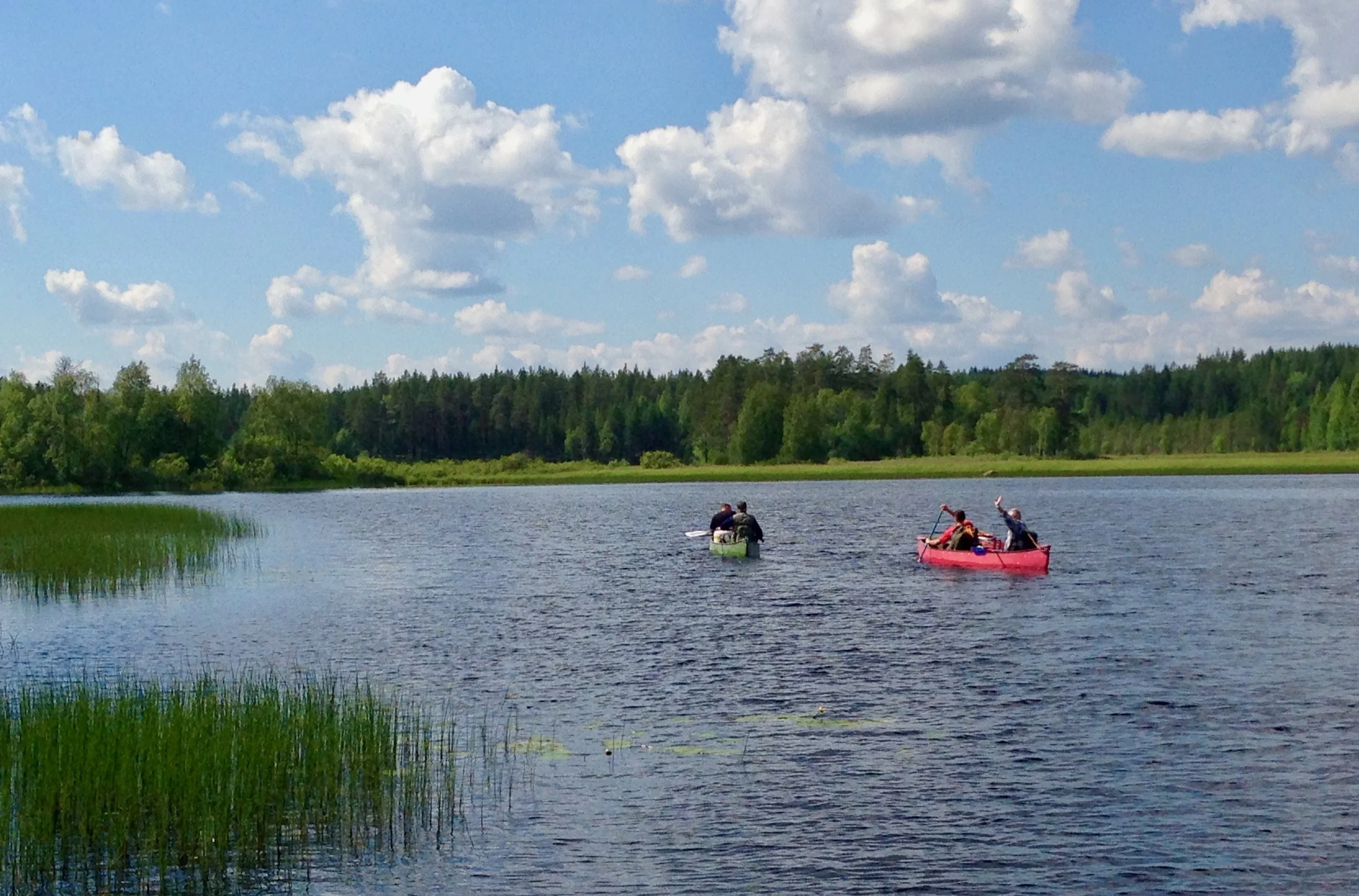 Two people kayaking on a lake with a forested shoreline, blue sky, and scattered clouds.