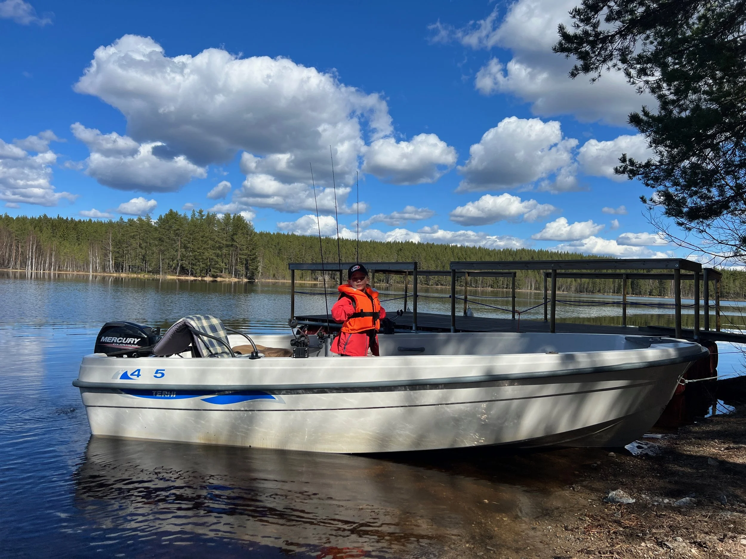 A child in a red jacket and orange life vest standing on a boat by a lake, holding fishing rods, with trees and a cloudy sky in the background.
