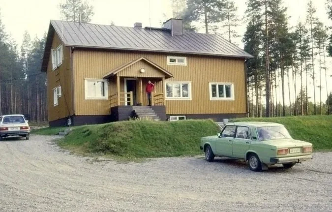 A two-story brown house with a metal roof, situated on a grassy hill, with cars parked in front and on the side. The house has a small porch with a person standing on it. Trees are visible in the background.