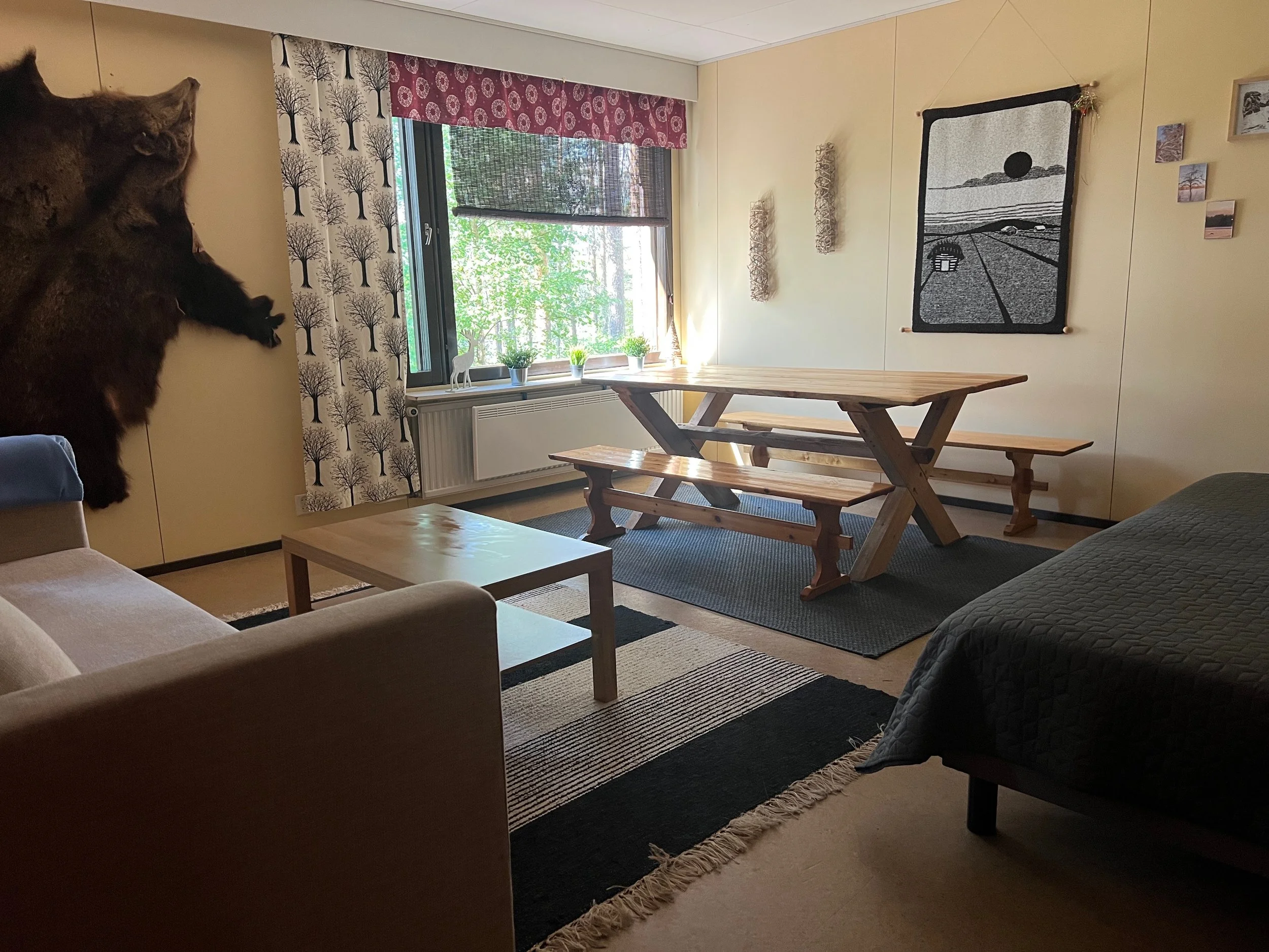 Living room Kokkojärvi with a window, a bench, a rug, wall decorations, and a large wall hanging artwork, with a sofa and a bed partially visible.