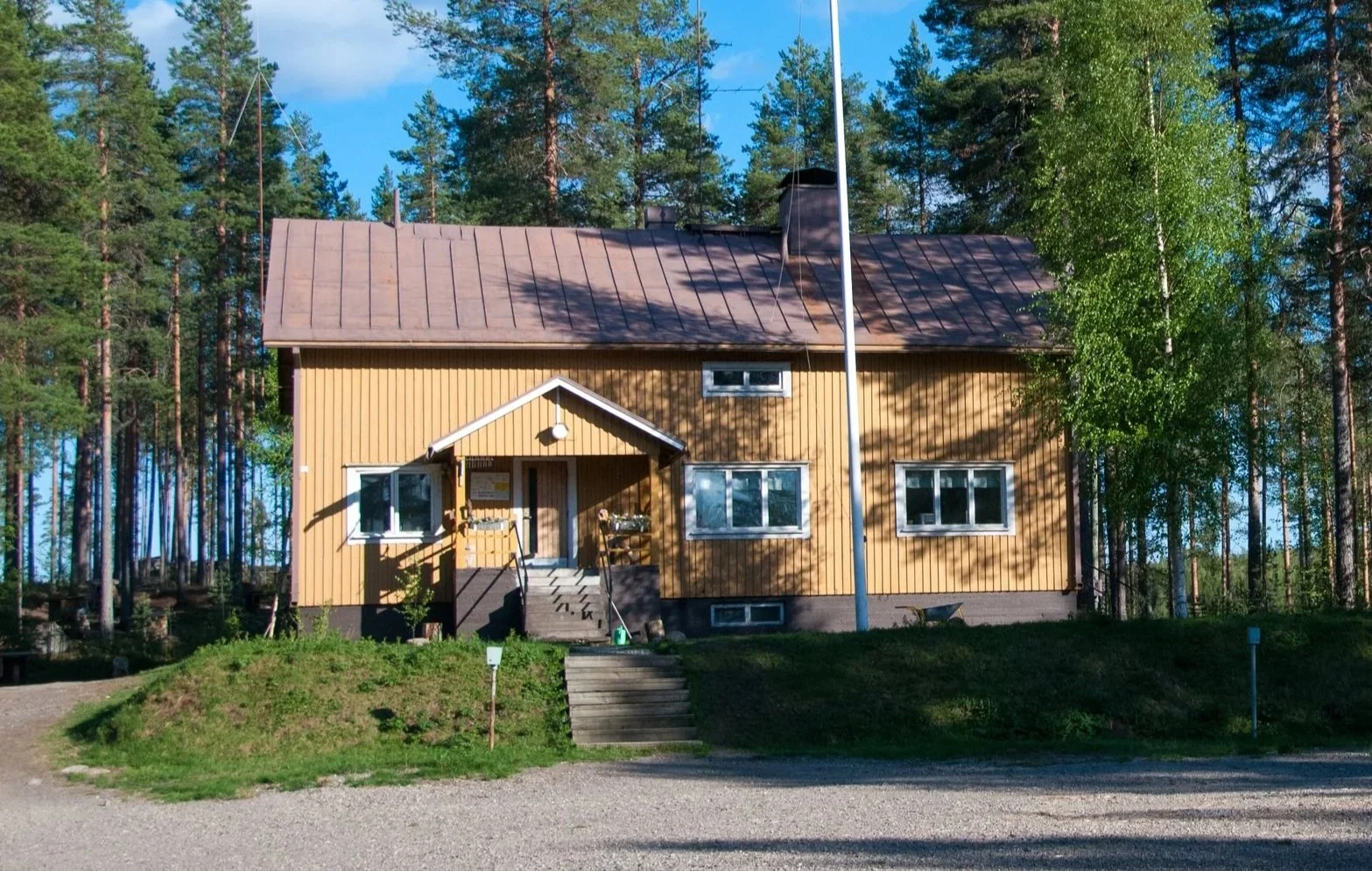 A yellow wooden house with white trim, set against a backdrop of tall pine trees and a bright blue sky. The house has a small front porch with stairs leading up to it and is surrounded by green grass and gravel.