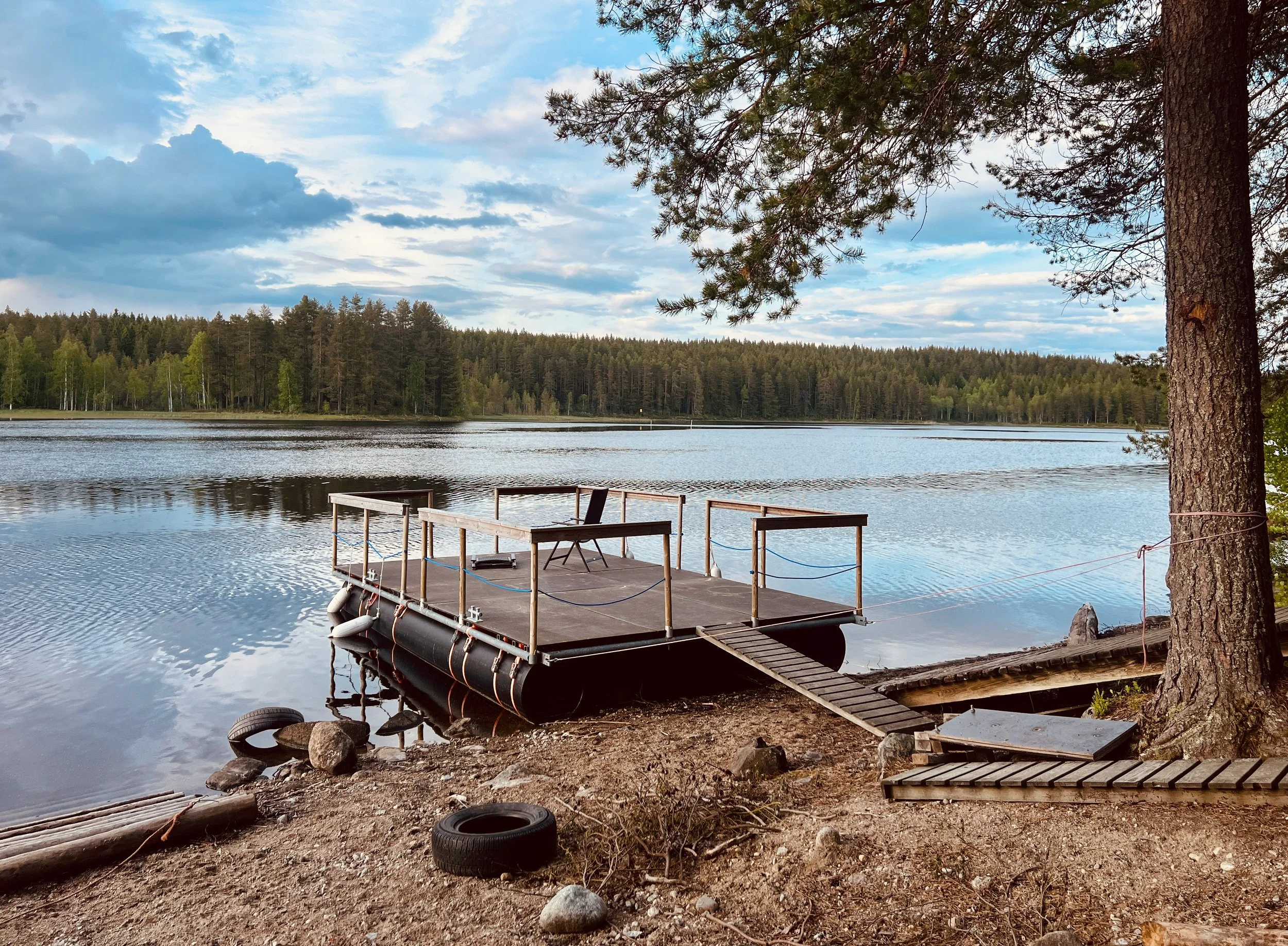A floating dock with a single chair extends into a calm lake surrounded by trees. The sky is partly cloudy with patches of blue.
