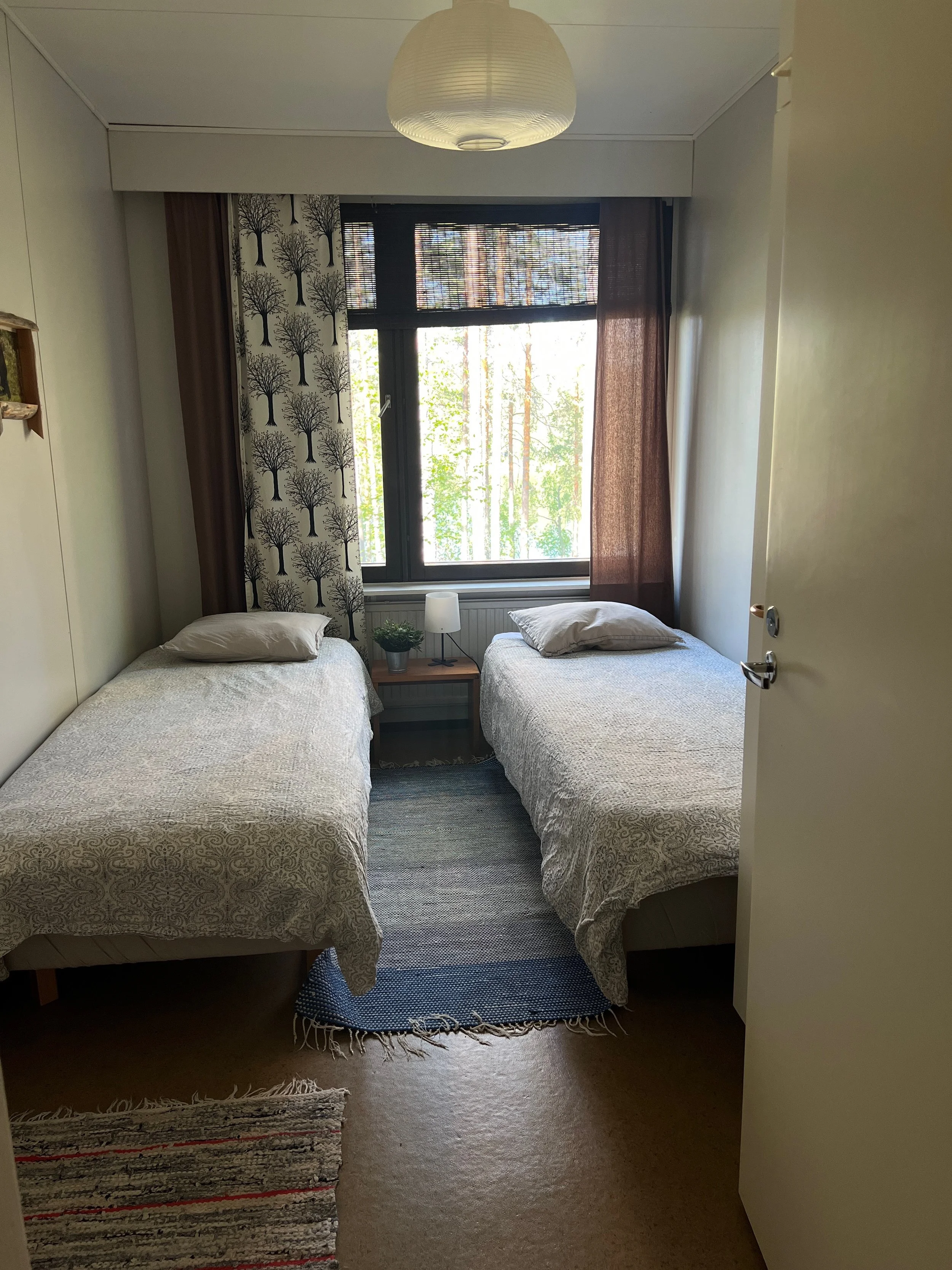 A bedroom with two single beds, a window with curtains, a nightstand with a lamp and plant between the beds, and a ceiling light fixture.