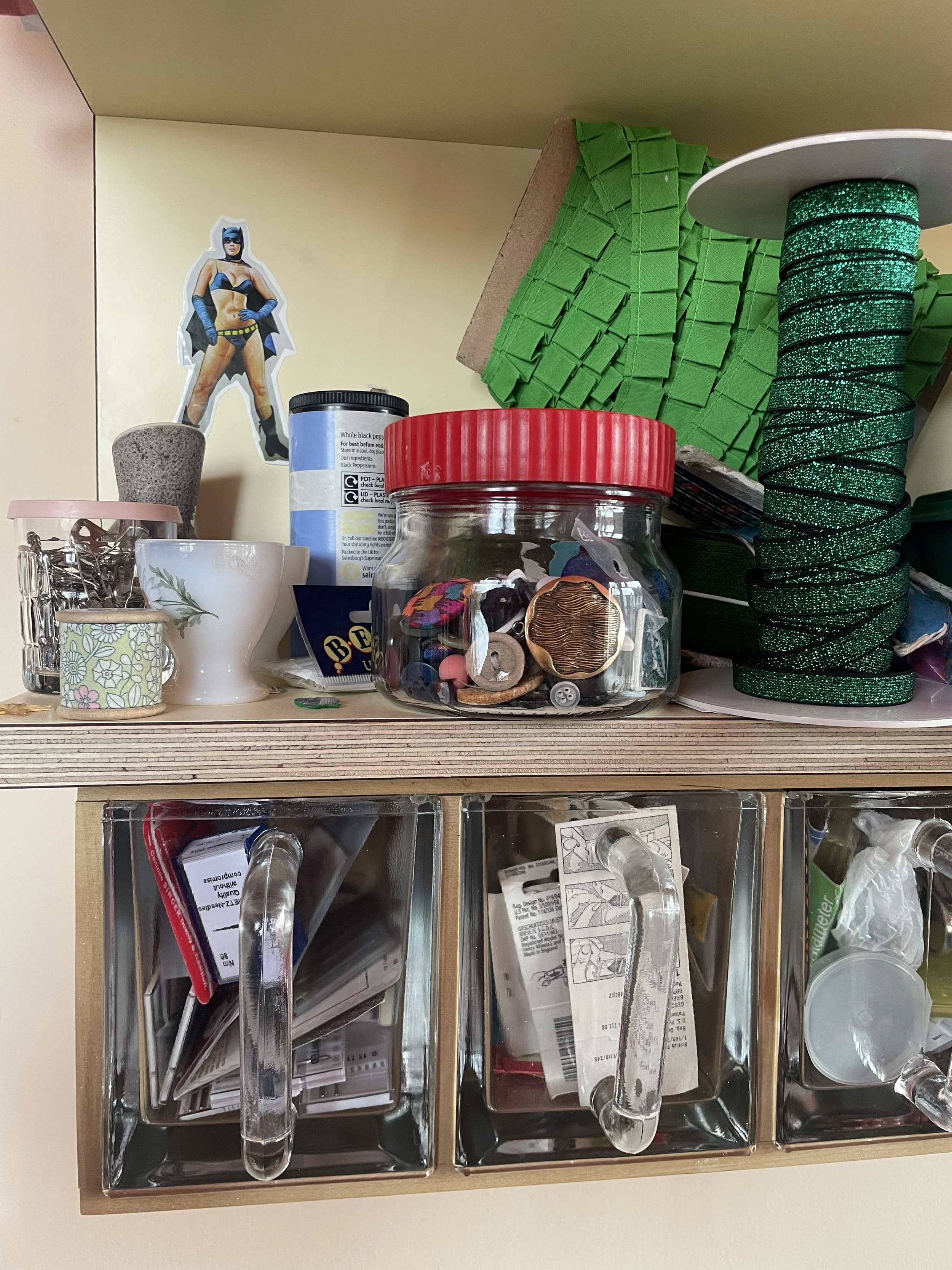 A wooden shelf with glass-front compartments containing various small items, above which is a cluttered surface with buttons in a jar, decorative cups, a cardboard piece, and green fabric trim, with a Batman figure sticker on the wall behind.