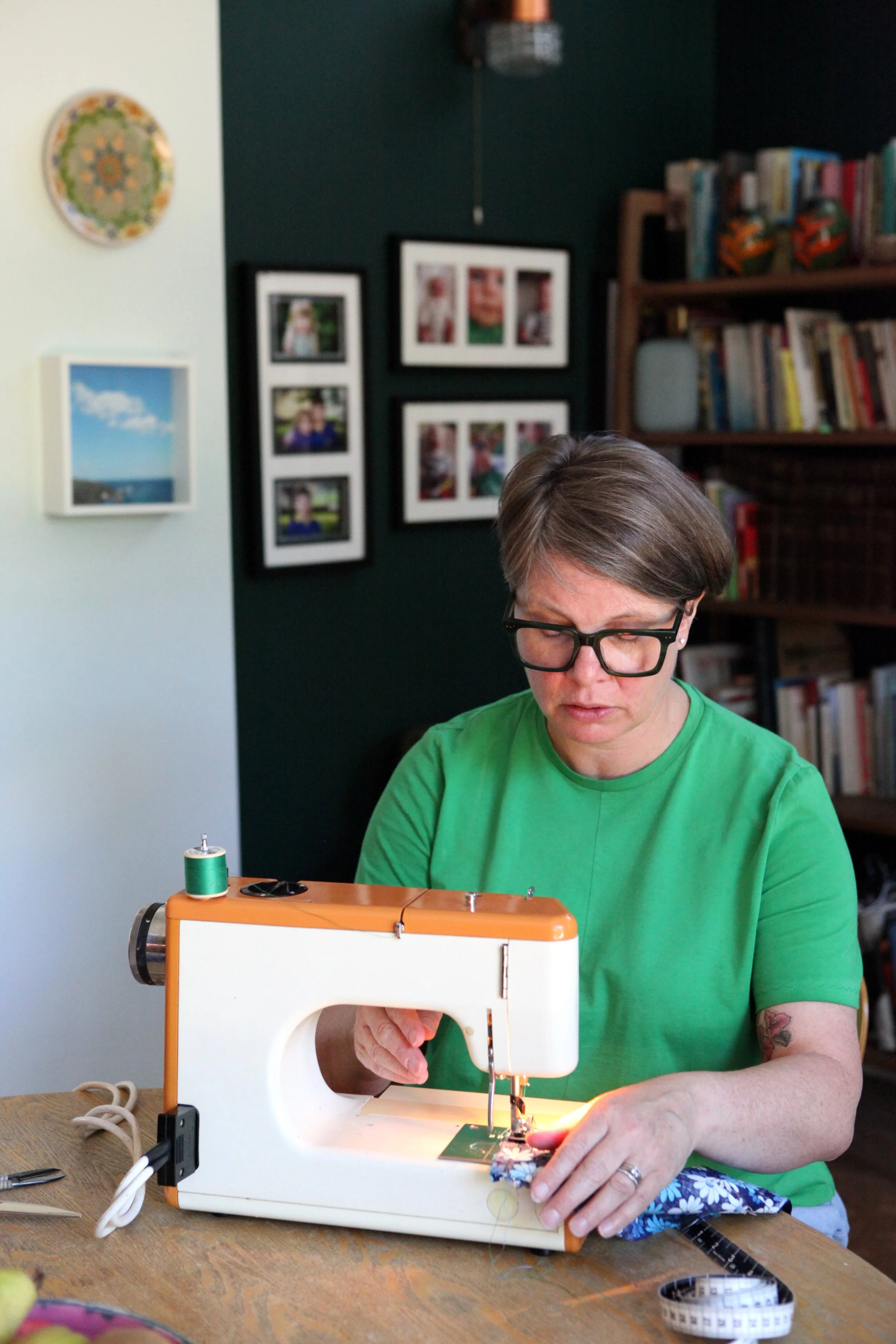 A woman wearing glasses and a green t-shirt is sewing fabric using a small sewing machine in a room with framed photographs and a bookshelf.