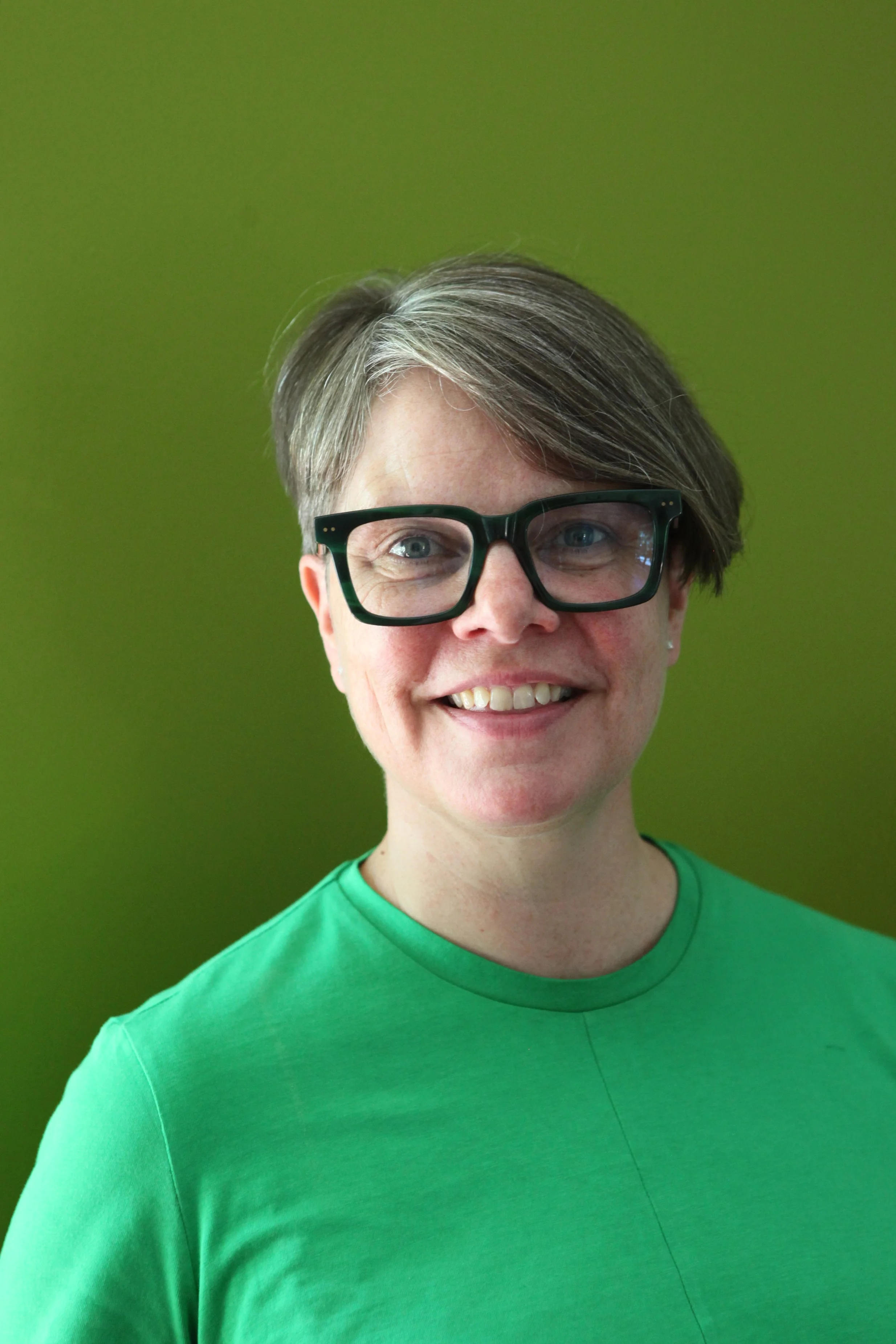 A woman with short gray hair and glasses smiling against a green background.