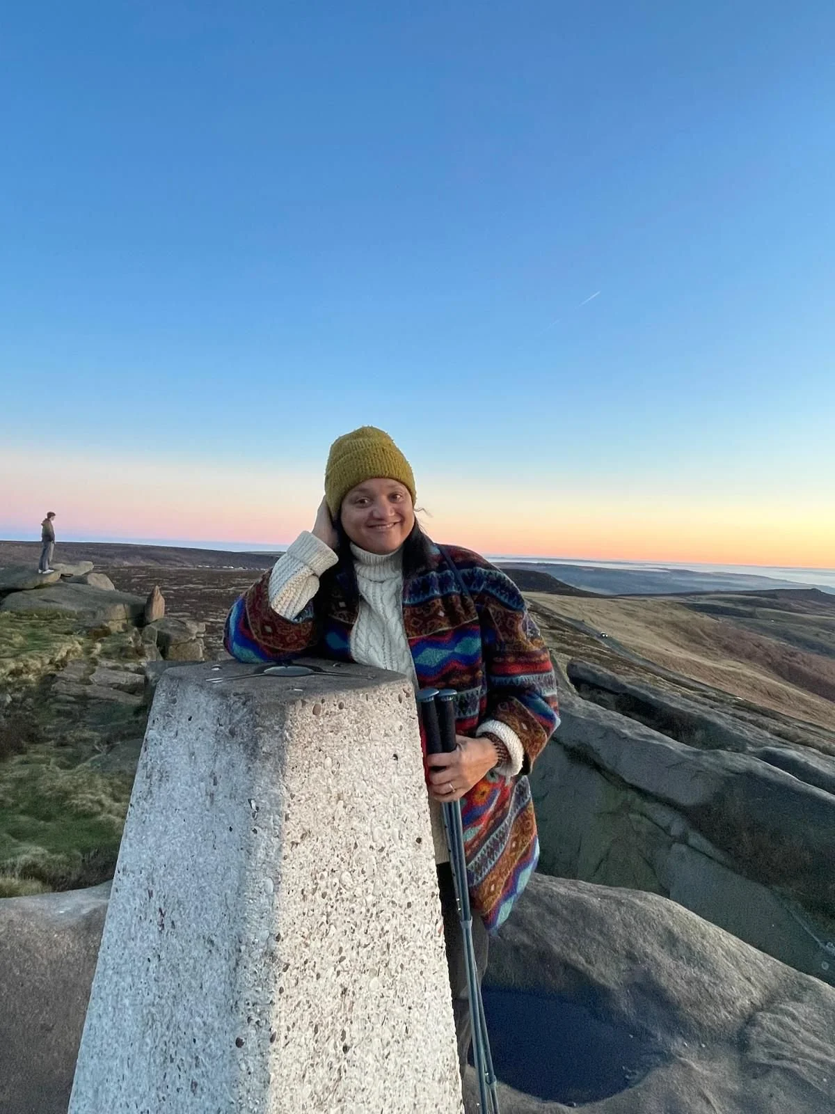A woman in a colorful patterned coat that is open and yellow knit hat leaning on a stone marker at sunrise or sunset in a hilly landscape, holding a walking cane. She is smiling.