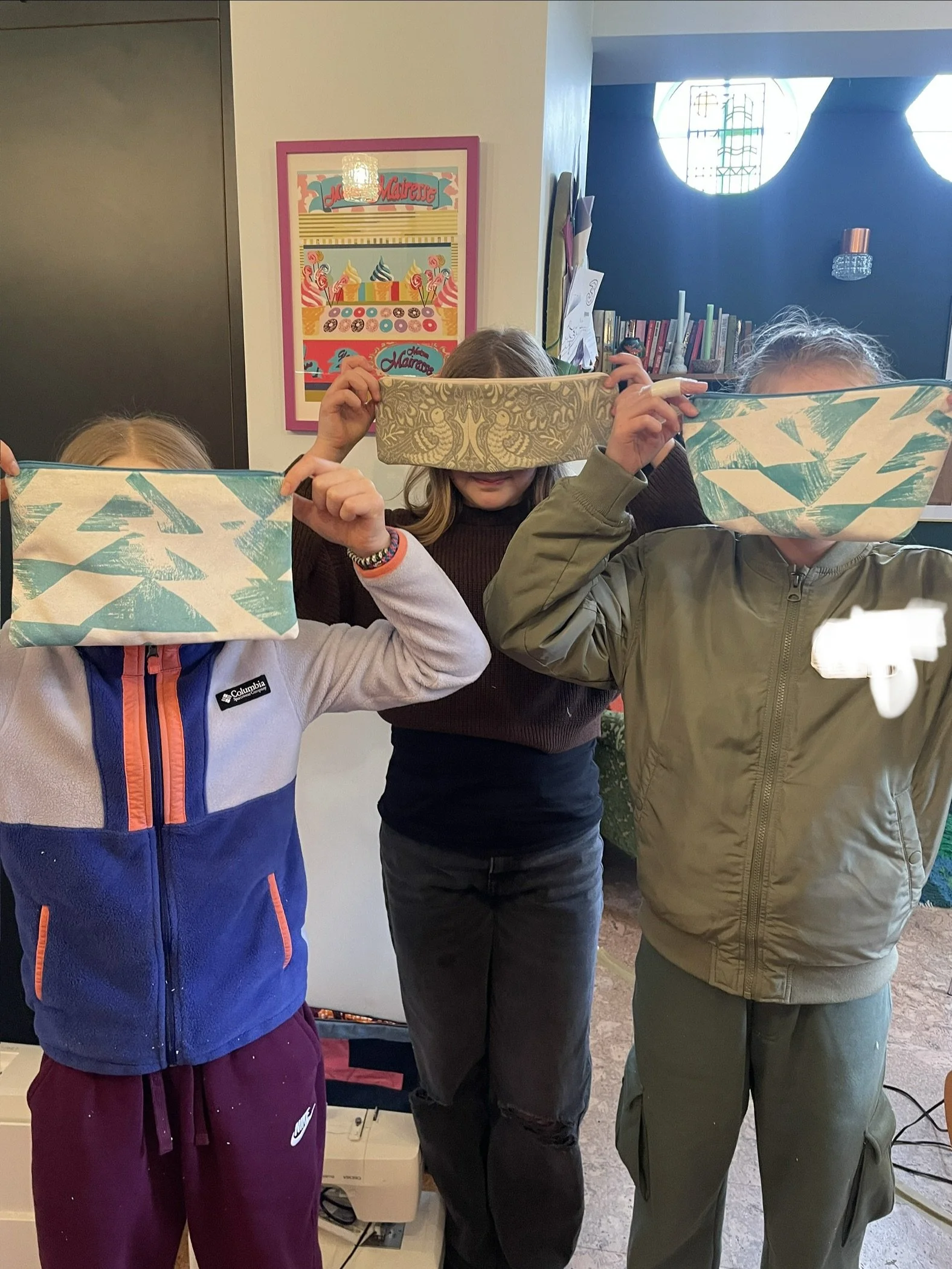 Three children are holding up patterned pencil cases in front of their faces, standing in an indoor setting with a colorful wall art and bookshelves in the background.