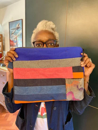 A person with short, curly gray hair and glasses is holding a large fabric bag in front of their face, partially covering it. The bag is made of pieced fabric in horizontal stripes of blue, pink, black, orange, and a multicolored pattern.