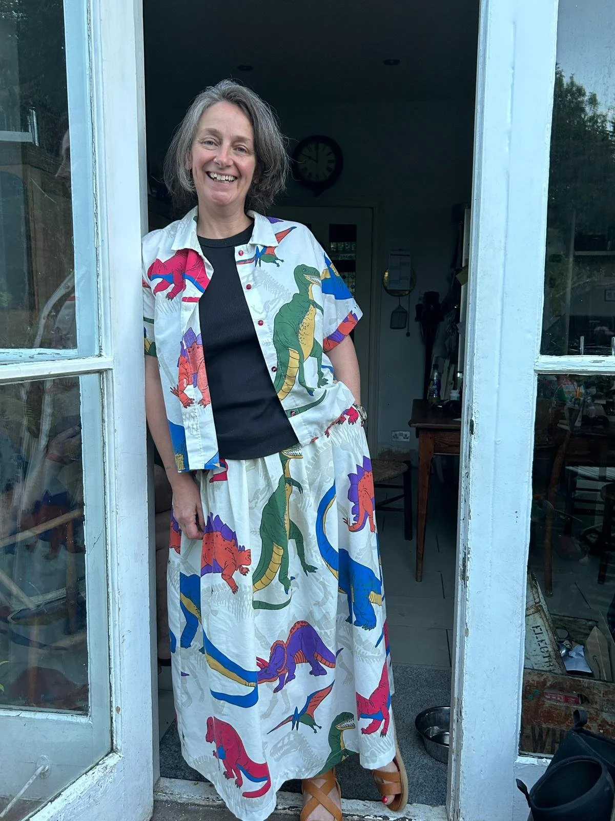 A woman standing at a doorframe, smiling, wearing a matching shirt and long skirt with colorful dinosaur prints.
