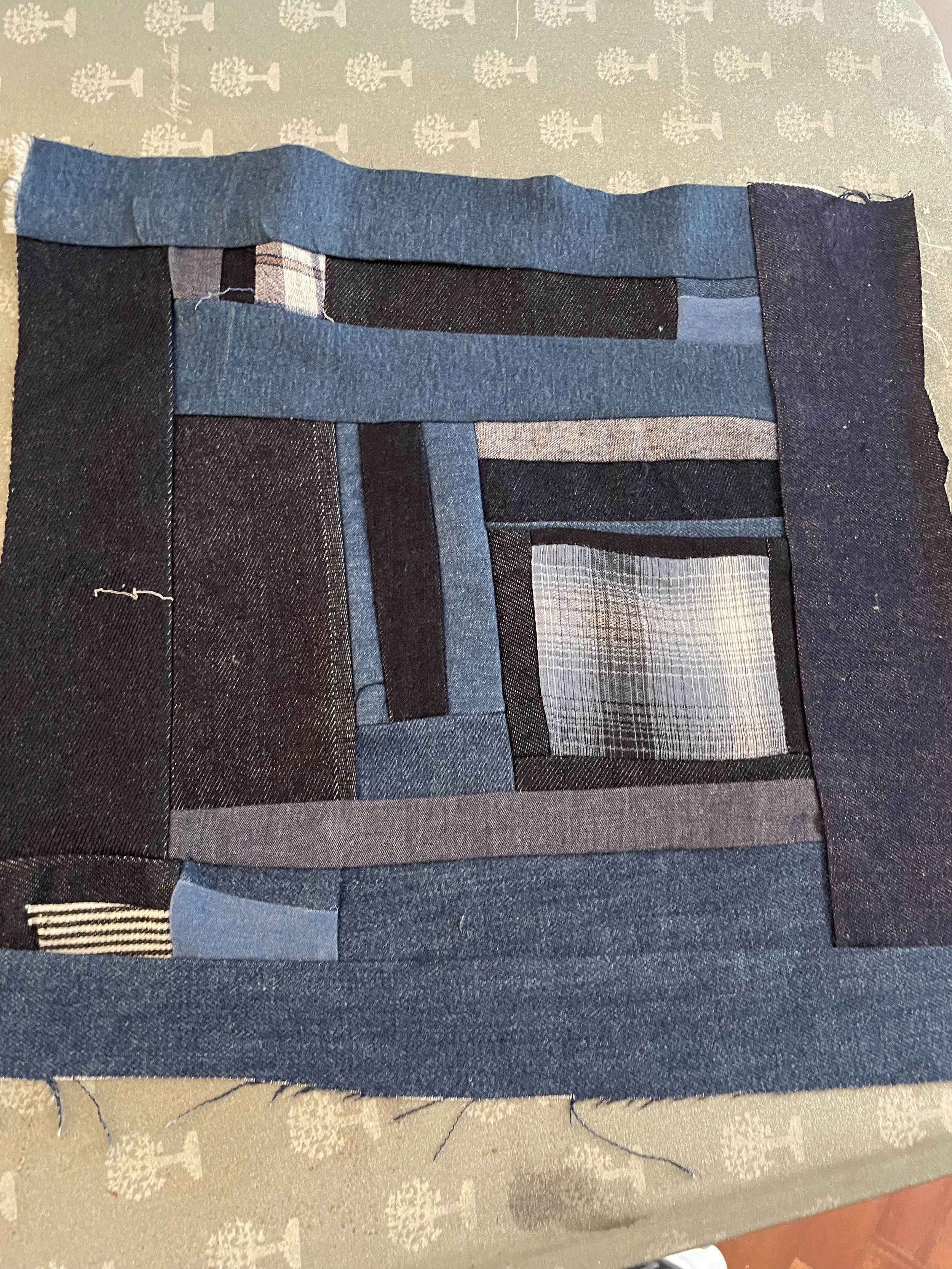 A fabric patchwork with various shades of blue and grey denim, arranged in rectangular and striped patterns, on a surface with a decorative tablecloth in the background.