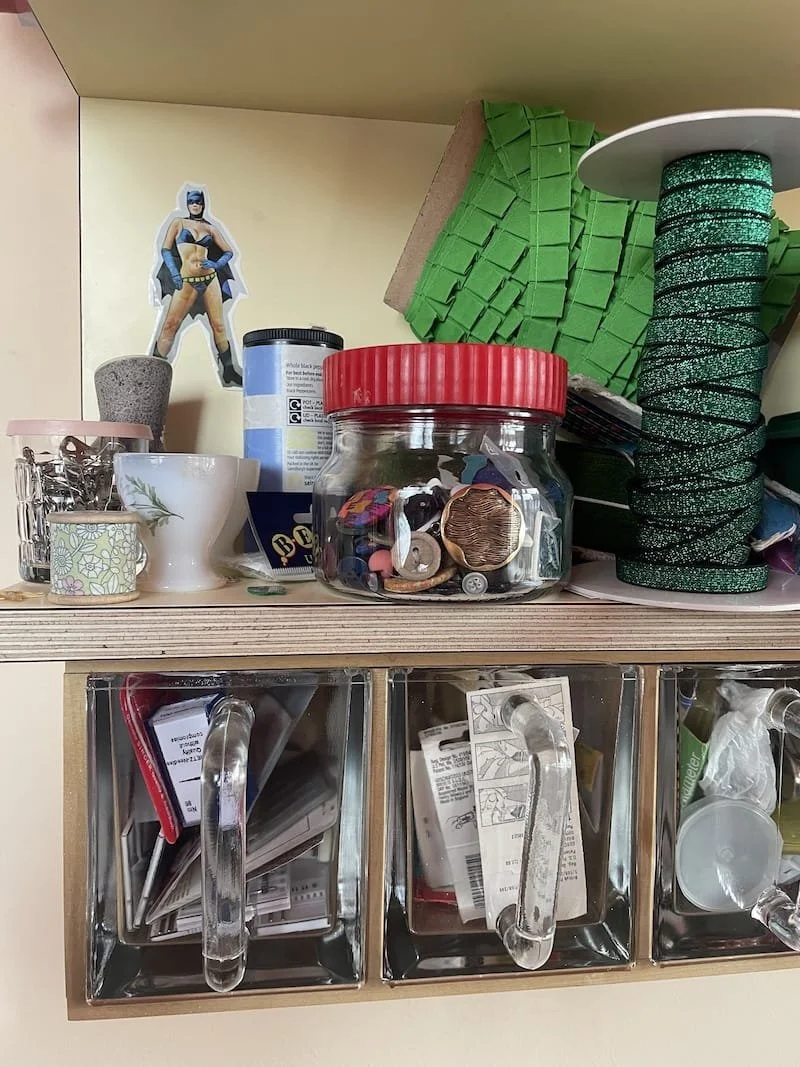 A pale yellow wooden shelving unit with glass fronted compartments. On the shelf are glass jars with buttons, rolls of elastic and green trim as well as small vessels with safety pins in. On the back wall of the shelf is a stick of batwoman