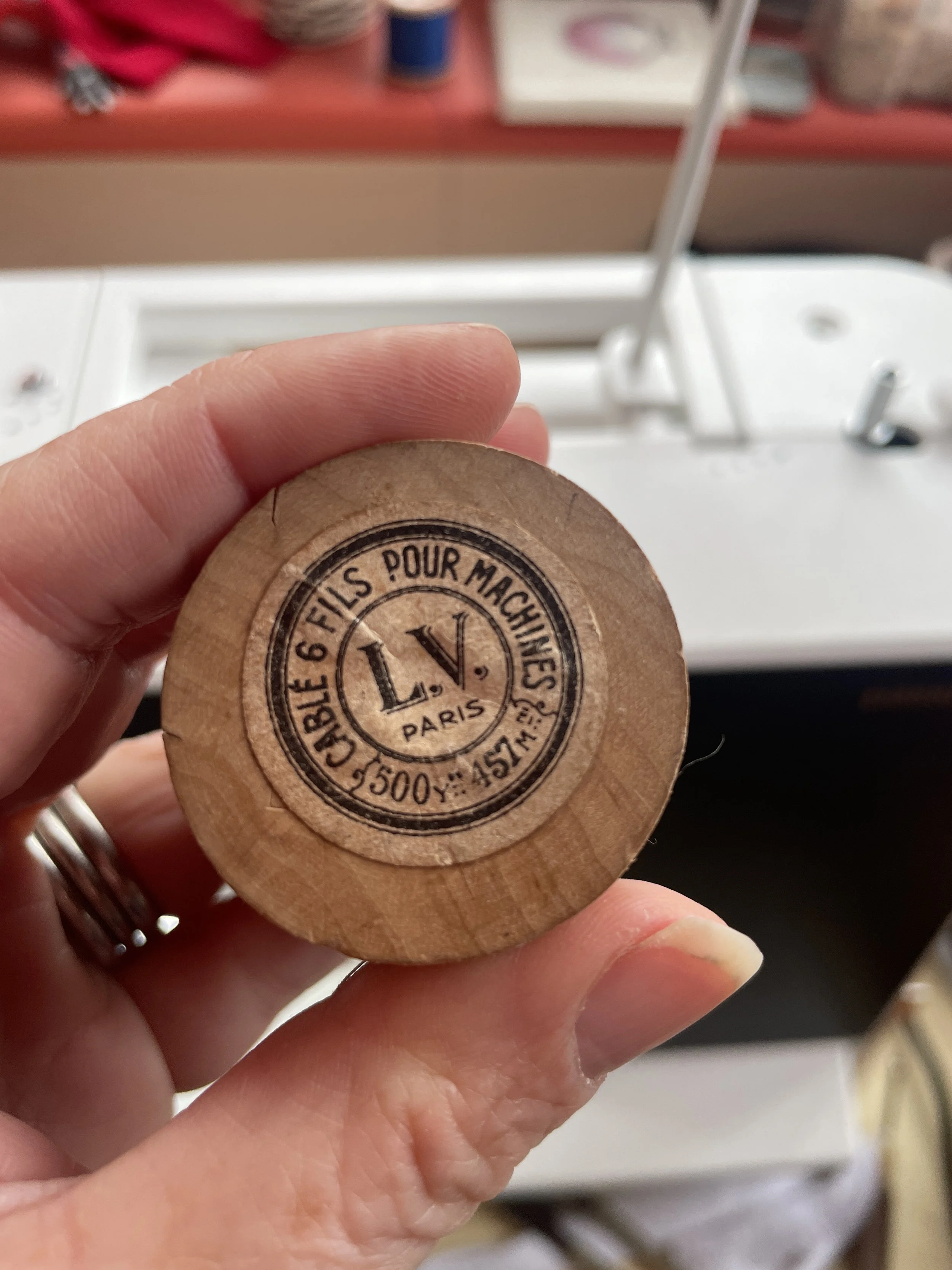 A person's hand holding a round wooden spool of thread with a Parisian stamp that reads, 'L.V. Paris, 500 Y, 154 M, Charle 6 Fils pour Machines'.