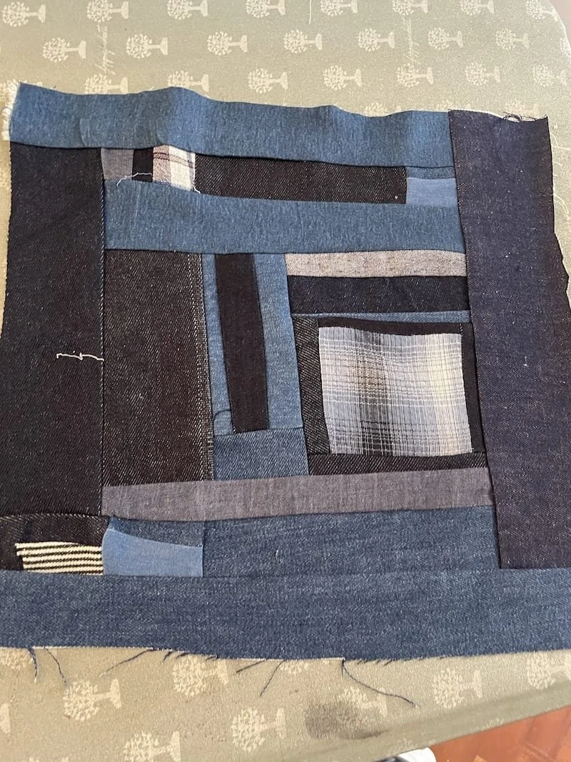 A fabric patchwork with various shades of blue and grey denim, arranged in rectangular and striped patterns, on a surface with a decorative tablecloth in the background.