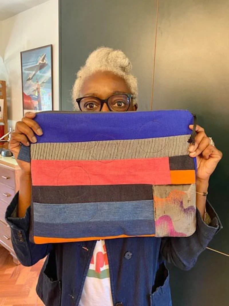 A person with short, curly gray hair and glasses is holding a large fabric bag in front of their face, partially covering it. The bag is made of pieced fabric in horizontal stripes of blue, pink, black, orange, and a multicolored pattern.