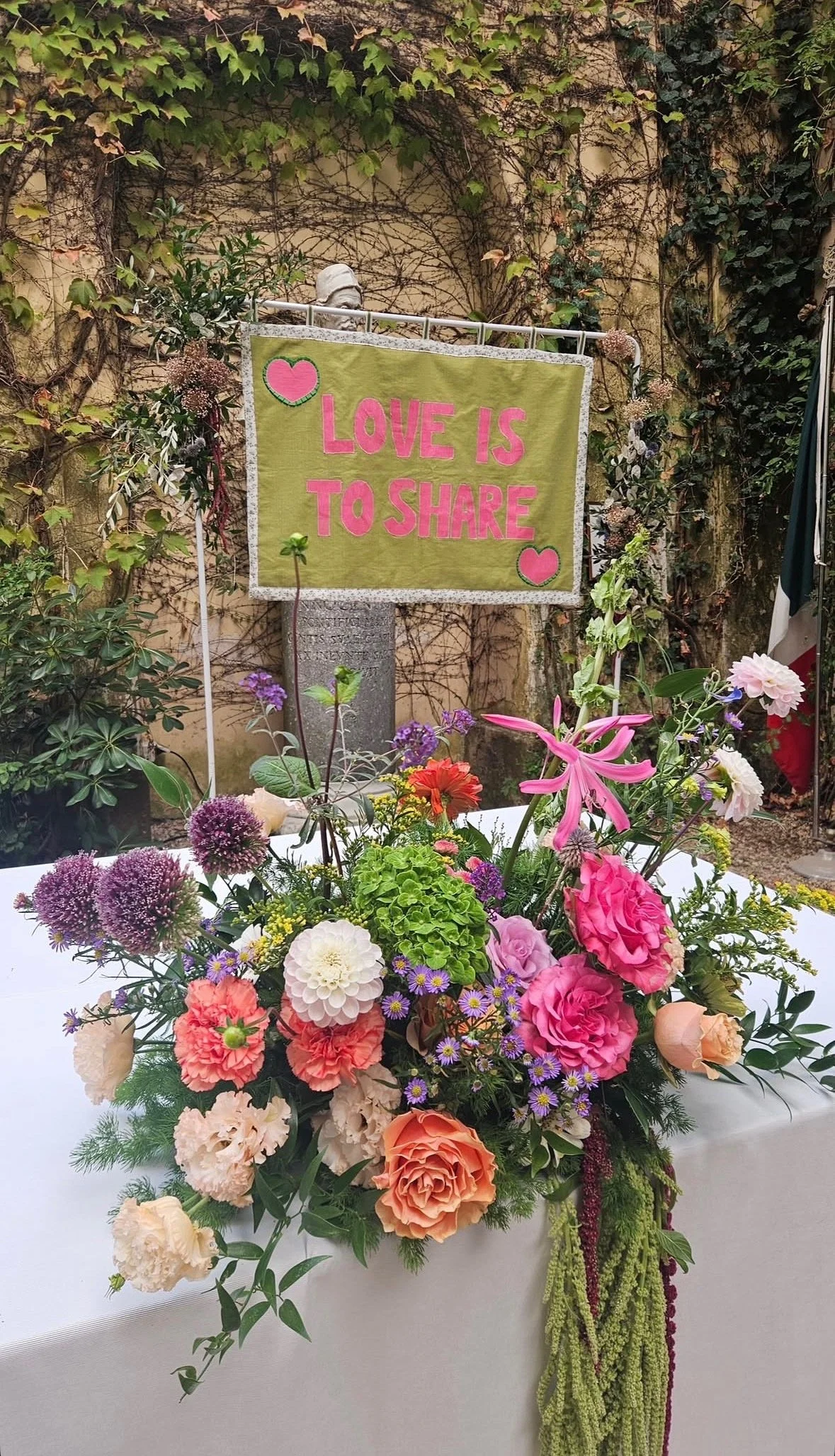 A colorful flower arrangement with pink lilies, roses, purple and white flowers, and greenery, situated on a white table. In the background, a sign reads 'Love is to share' in pink letters on a green fabric background, decorated with pink hearts. The