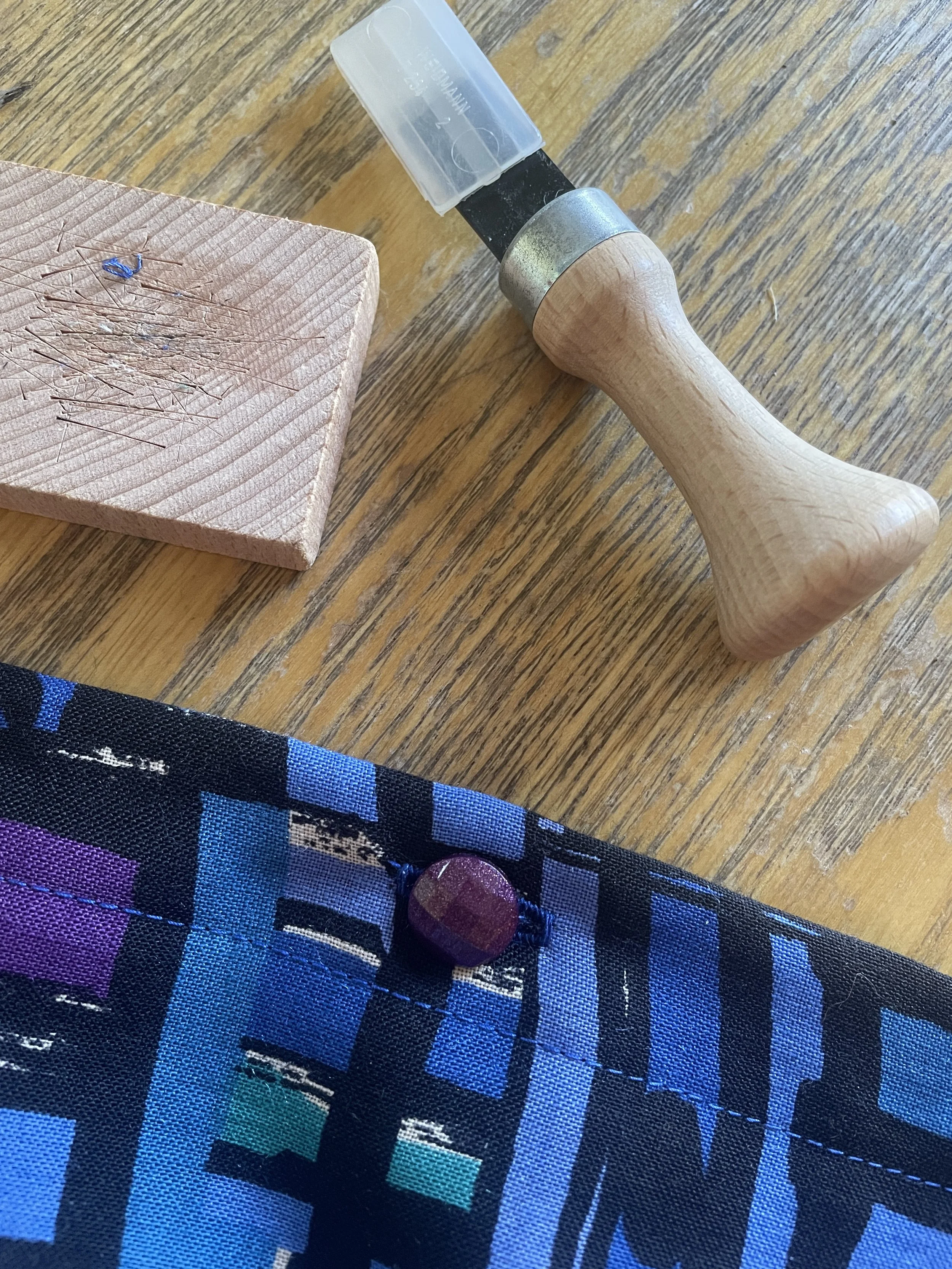 A wool fabric in various shades of dark blues and pink with a small purple faceted button sitting on a wooden table with a button chisel and block of wood 