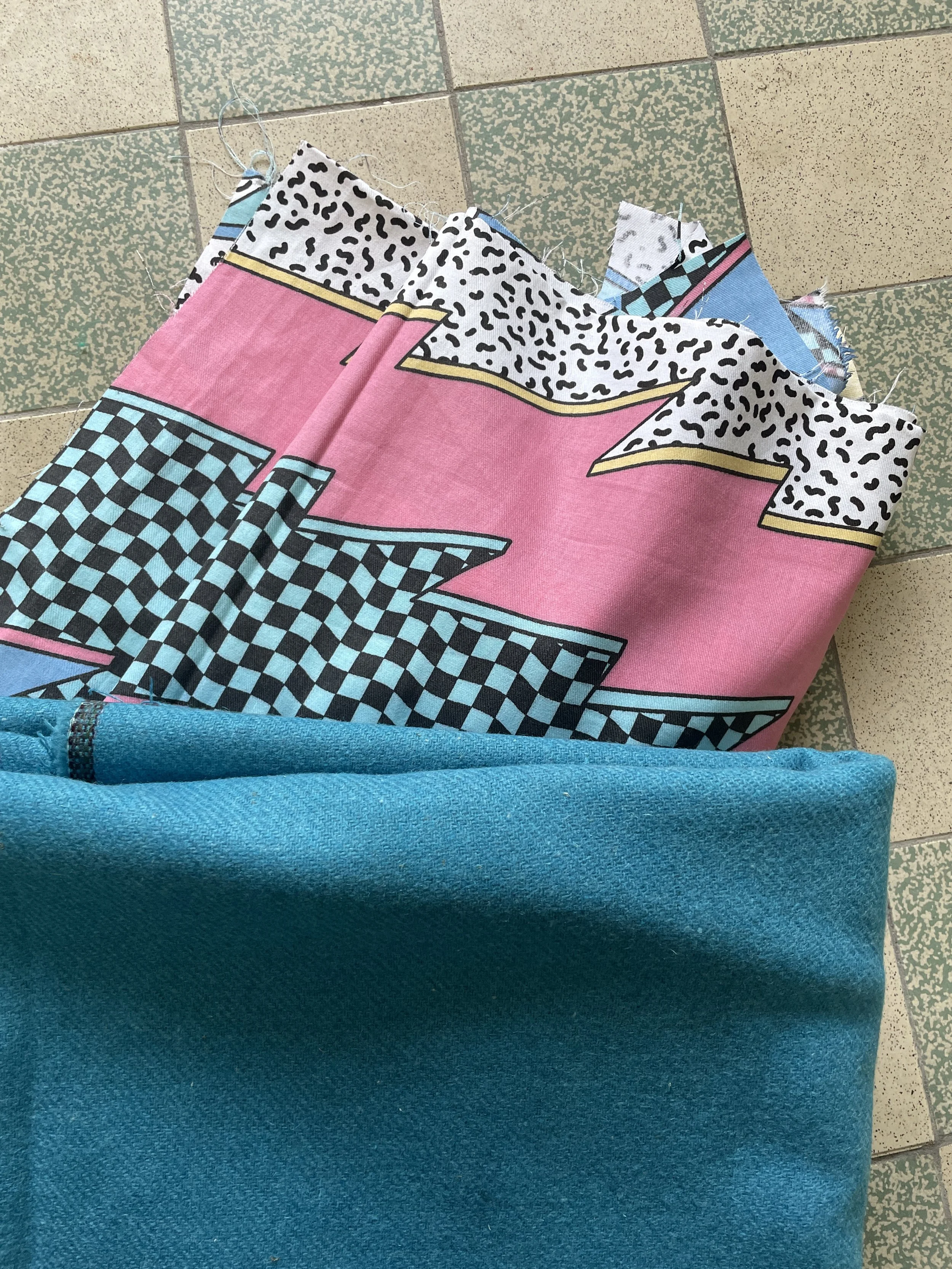 Colorful fabric with geometric patterns on the floor, underneath a turquoise wool fabric. 