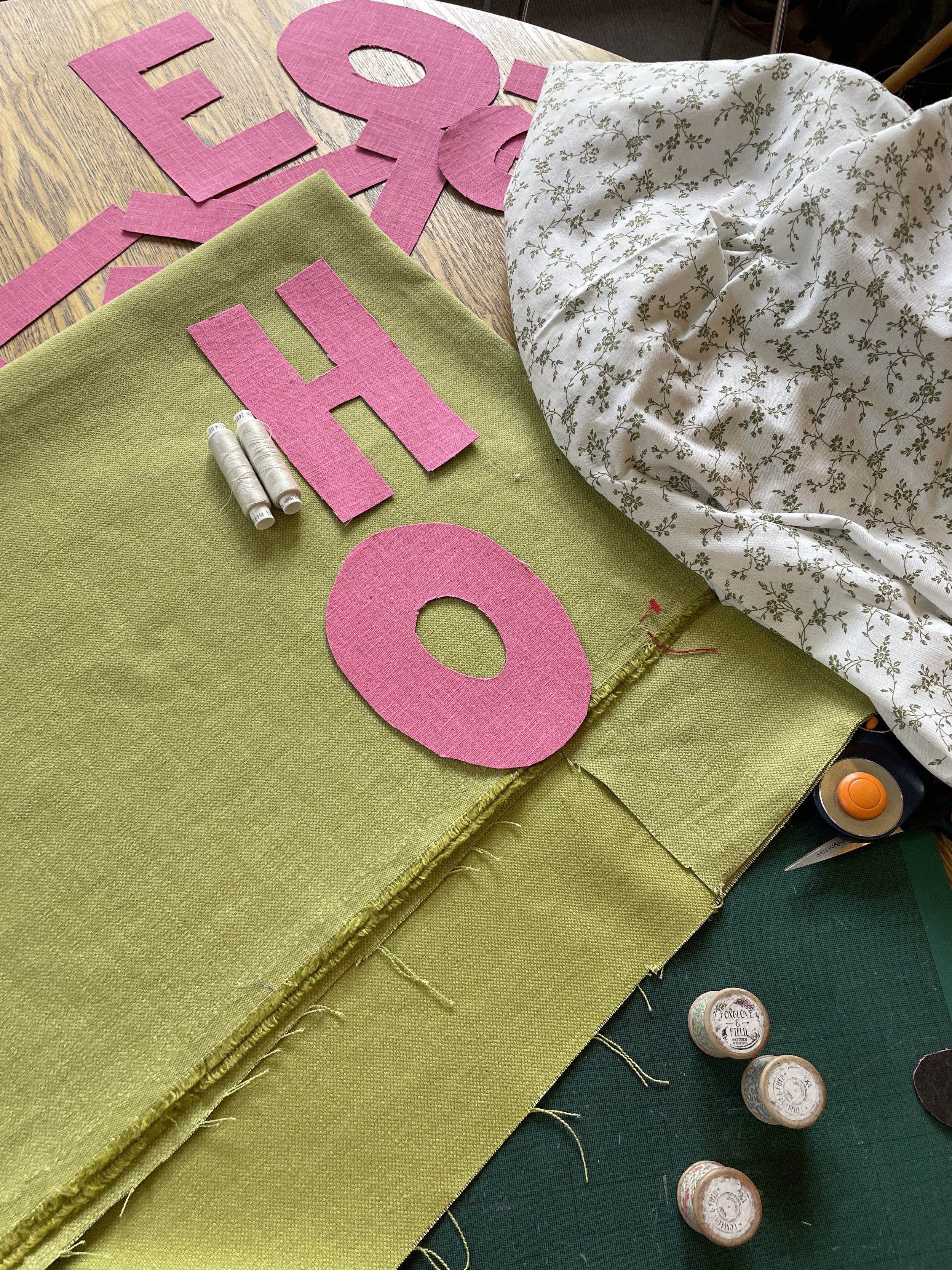 Crafting supplies including pink fabric letters spelling 'LOVE' and a large pink letter 'H' on green fabric, sewing threads, scissors, and white floral-patterned fabric on a worktable.