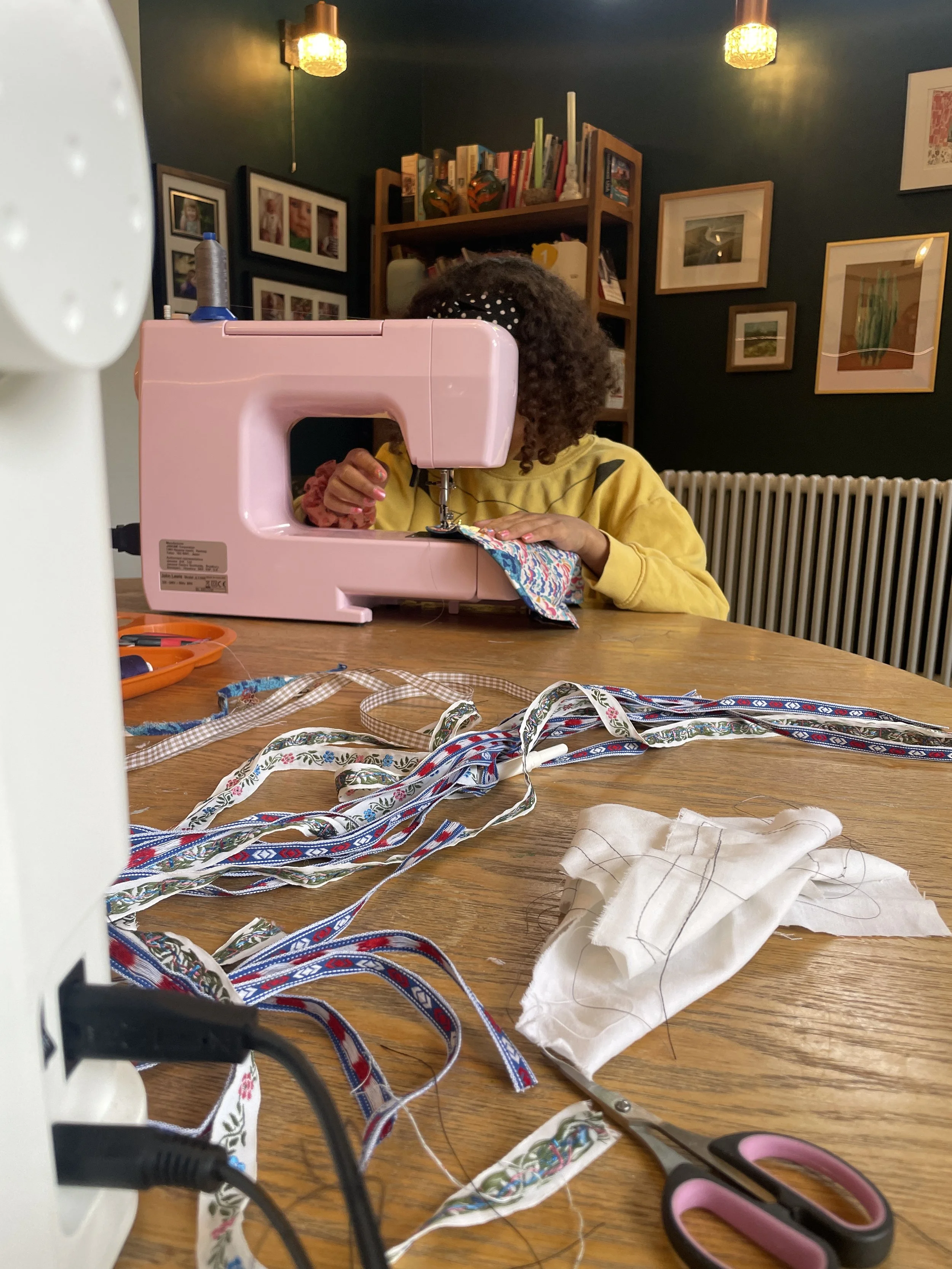 A girl sewing fabric on a pink sewing machine at a wooden table. The table has colorful ribbons, a pair of scissors, and fabric pieces. The background shows a wall with framed pictures and a bookshelf, with warm lighting overhead.