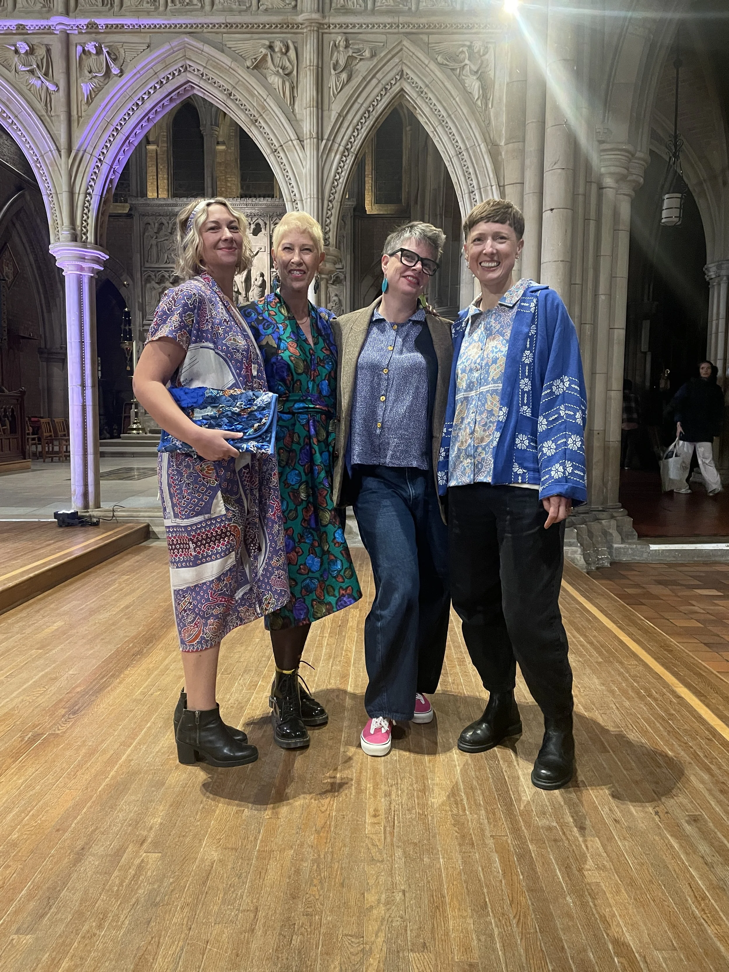 Four women standing together in a church with gothic architecture, smiling at the camera, wearing colorful outfits and black boots.