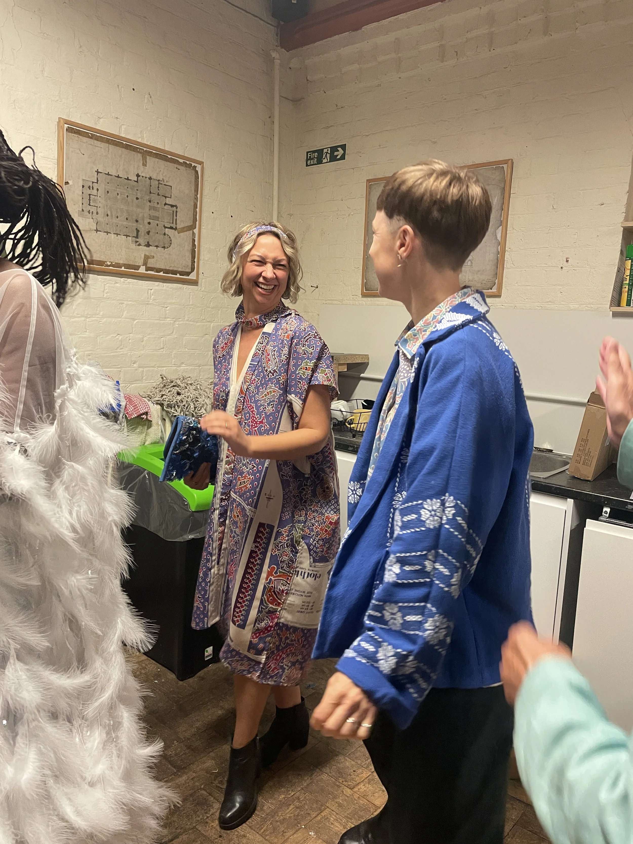 Two women are laughing and dancing at a social gathering in a room with white brick walls and decor.