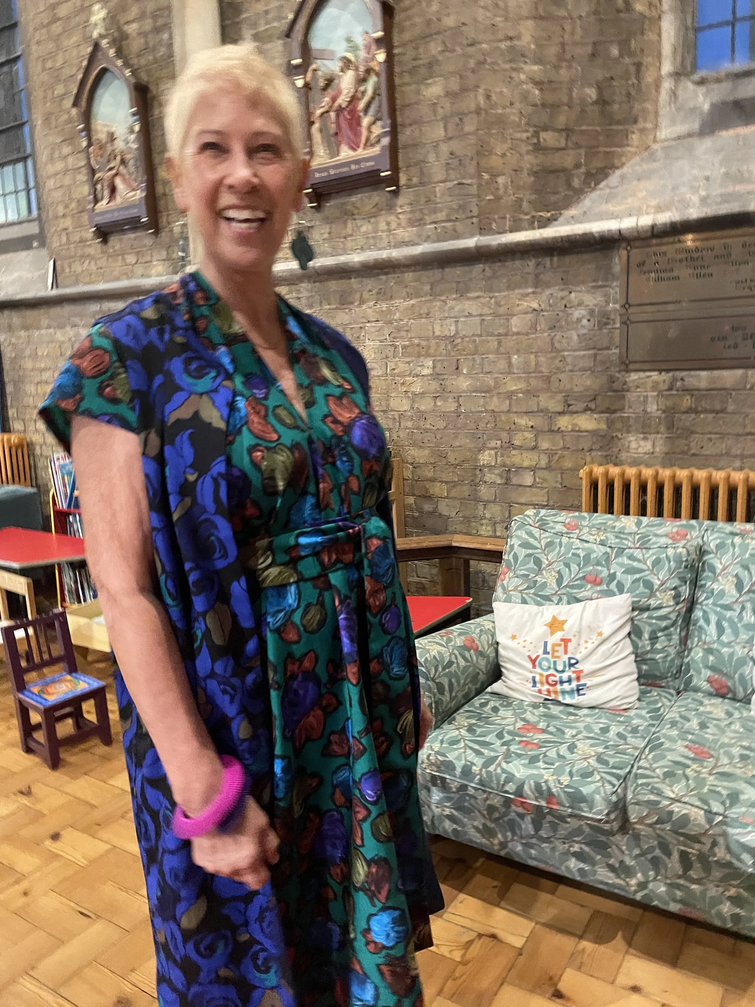 A woman with short blonde hair smiling, wearing a floral dress and long waistcoat and large pink bracelet, standing indoors against a brick wall
