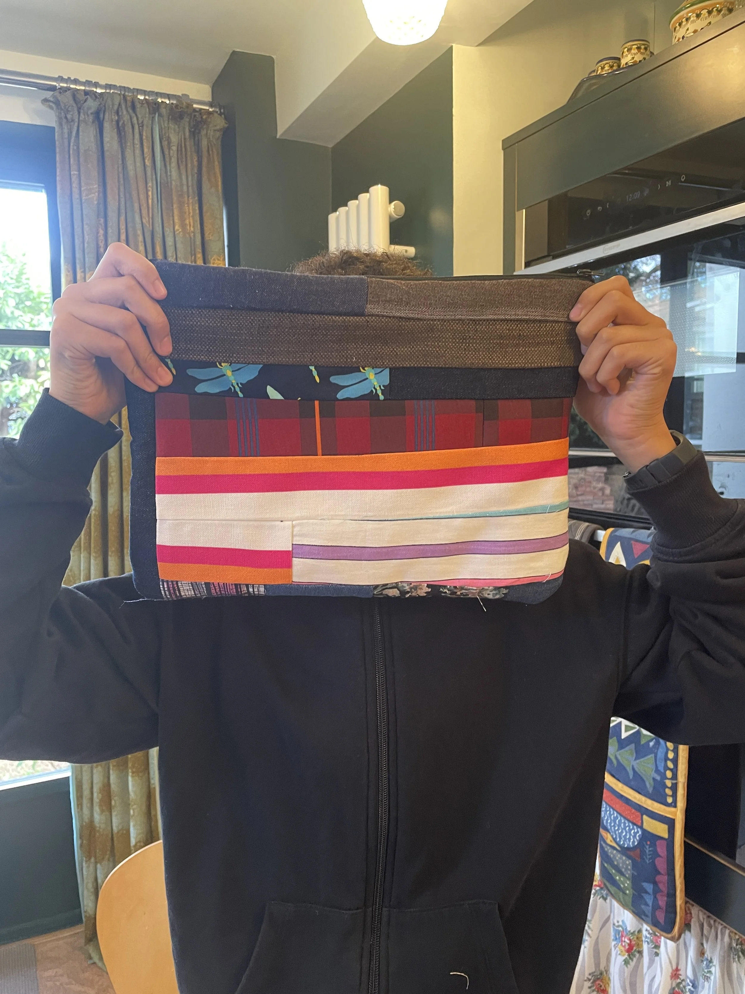 Person holding a fabric storage box with a colorful striped pattern in front of their face, in a kitchen with dark green walls, a window with floral curtains, a radiator, and kitchen appliances.