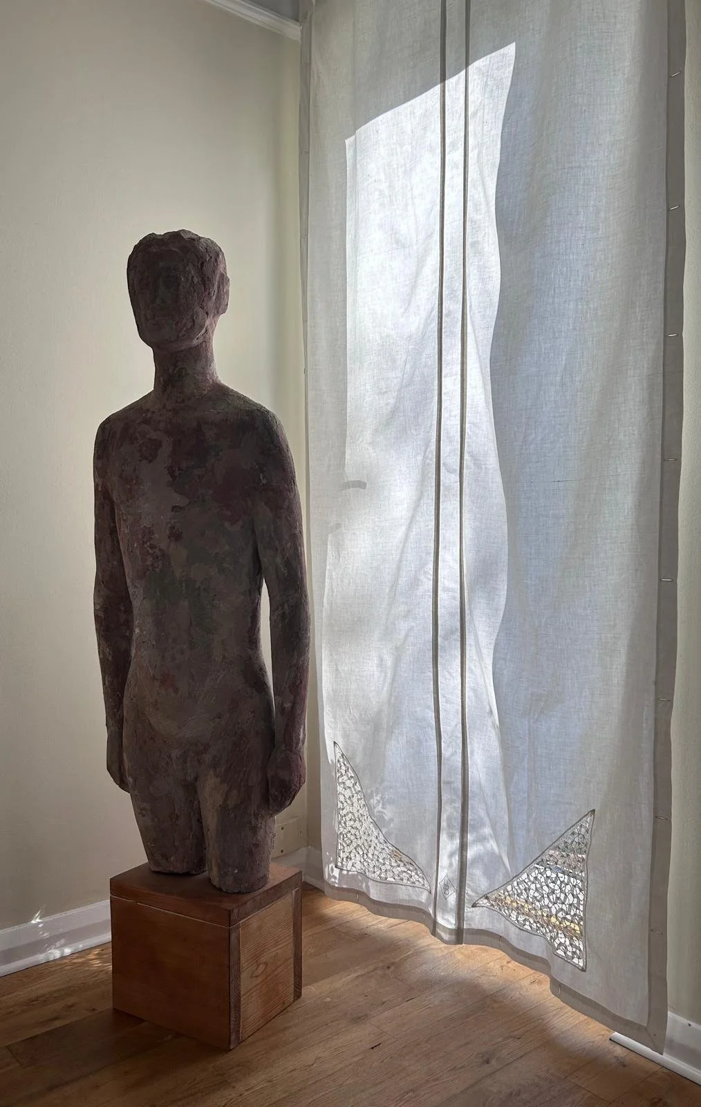 A sculpture of a person, unfinished and rough, standing on a wooden pedestal beside a white curtain with lace detailing, illuminated by natural light coming through and casting shadows.
