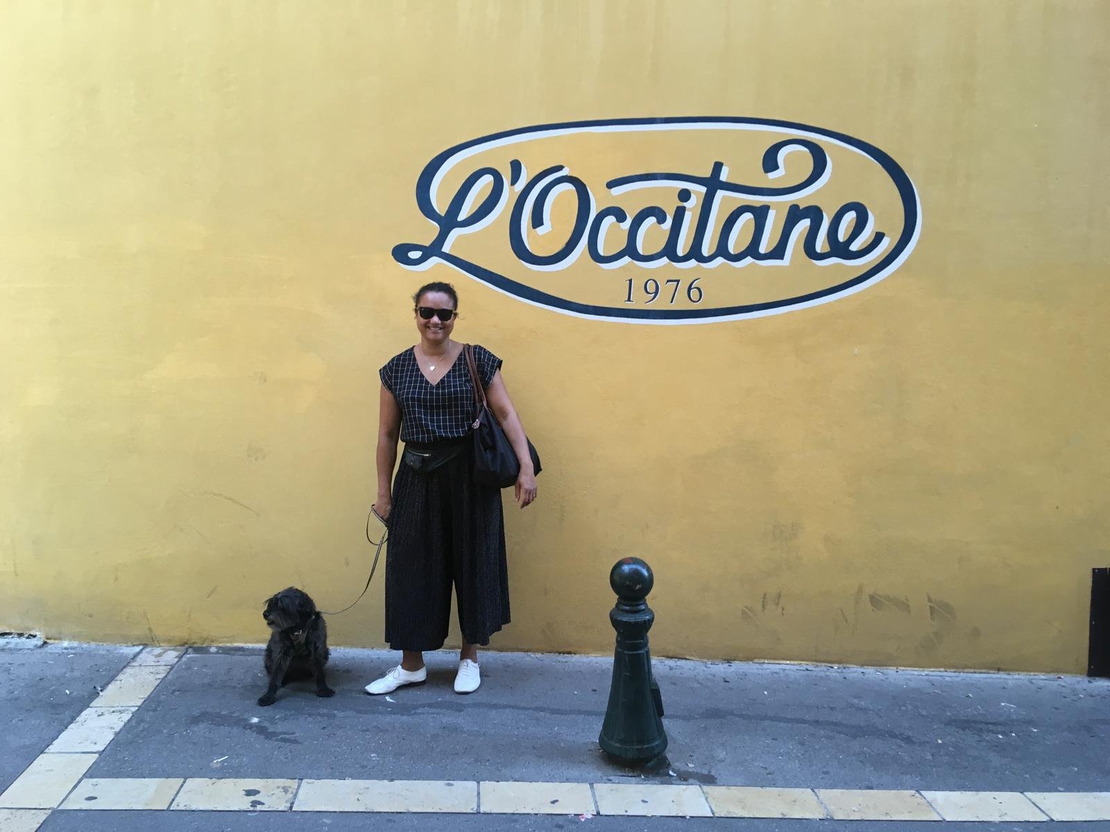 A woman with sunglasses standing on a street with a black dog on a leash. They are in front of a yellow wall with the sign 'L'Occitane 1976' painted on it.