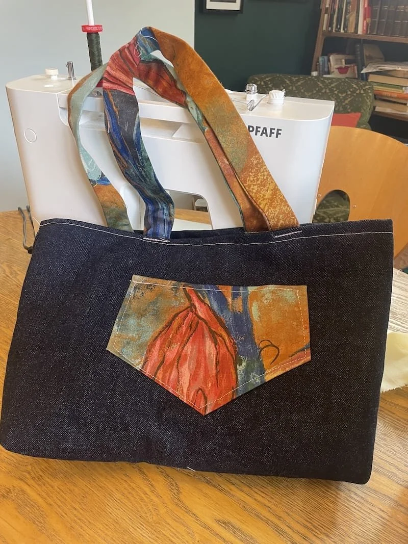 A black fabric tote bag with a colorful abstract-patterned pocket and handles, placed on a wooden table. In the background, there is a white sewing machine and bookshelf.