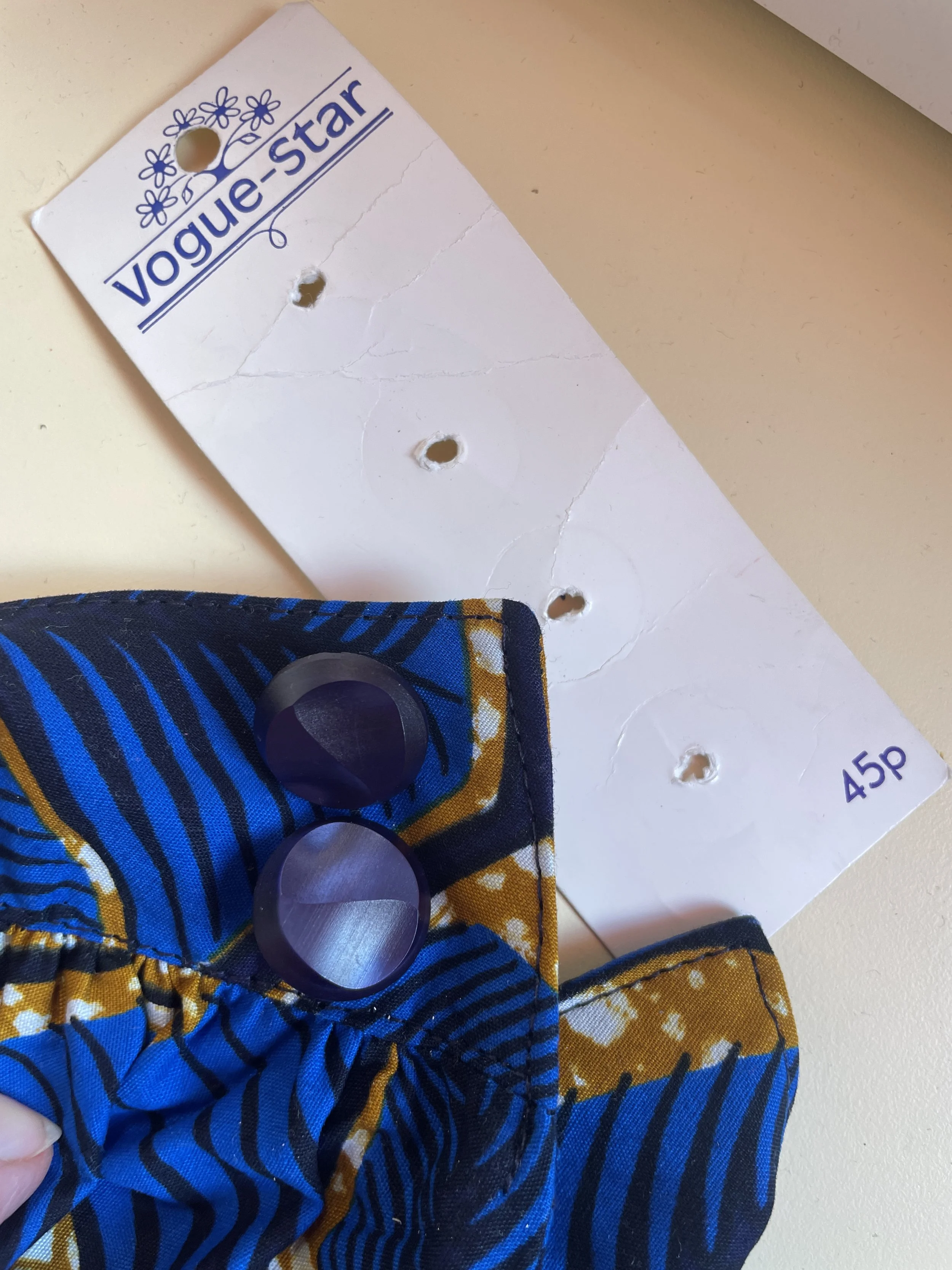 An vintage display card from a Vogue Star product that cost 45p with multiple holes punched through it that used to hold buttons, and two electric blue buttons sewn onto a blue and yellow patterned fabric, on a light-colored surface.