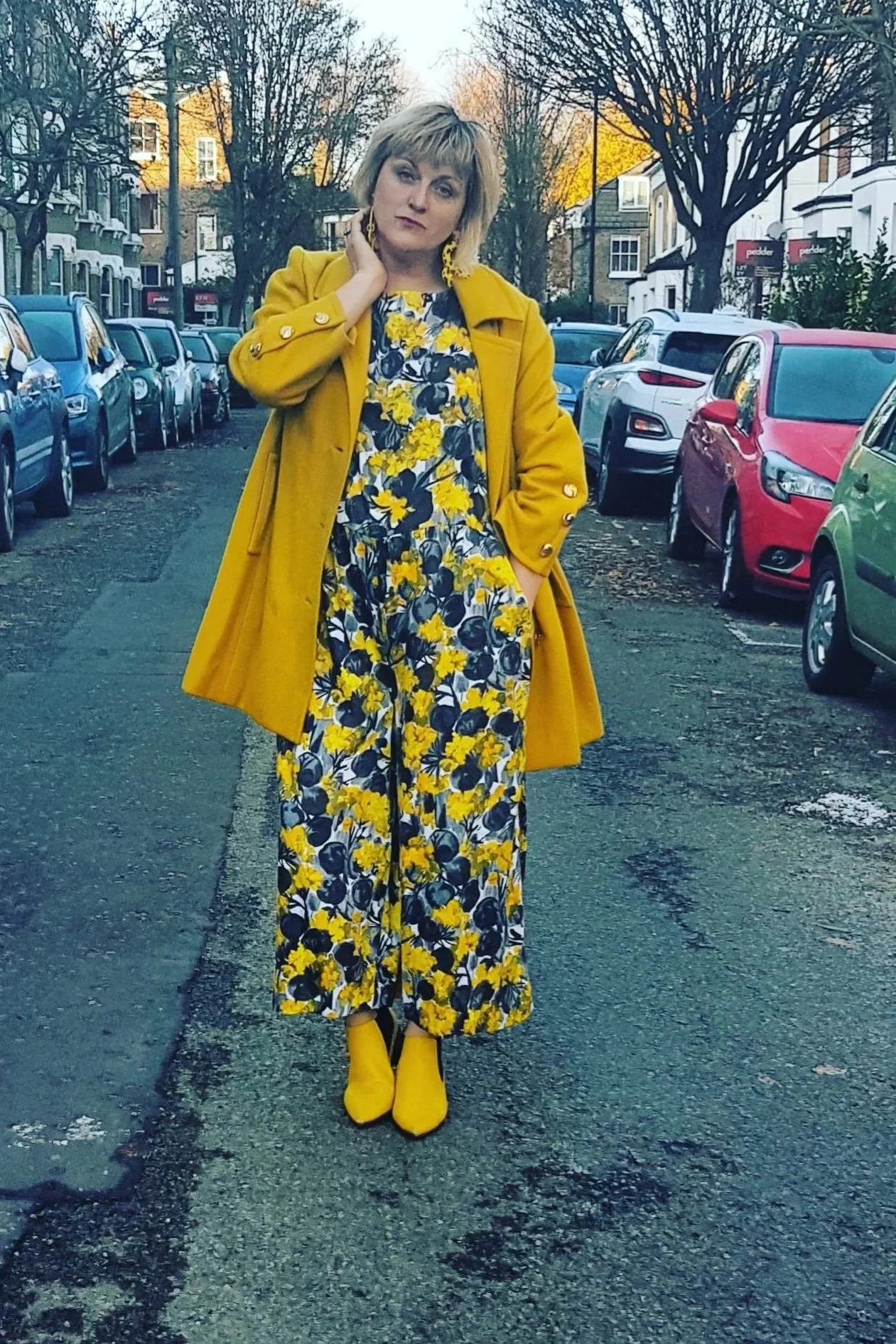Woman in a yellow coat and floral jumpsuit standing on a city street with parked cars and leafless trees.