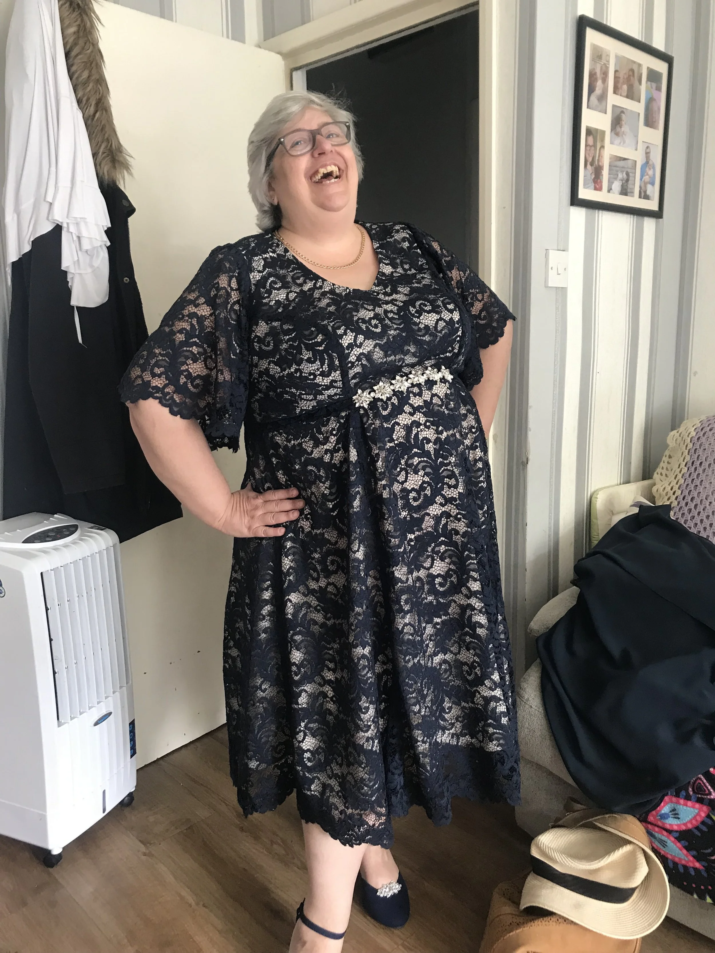 A woman dressed in a navy lace dress with floral embellishments, smiling and standing indoors in a room with wooden flooring, a picture frame on the wall, and various personal items including hats and bags.