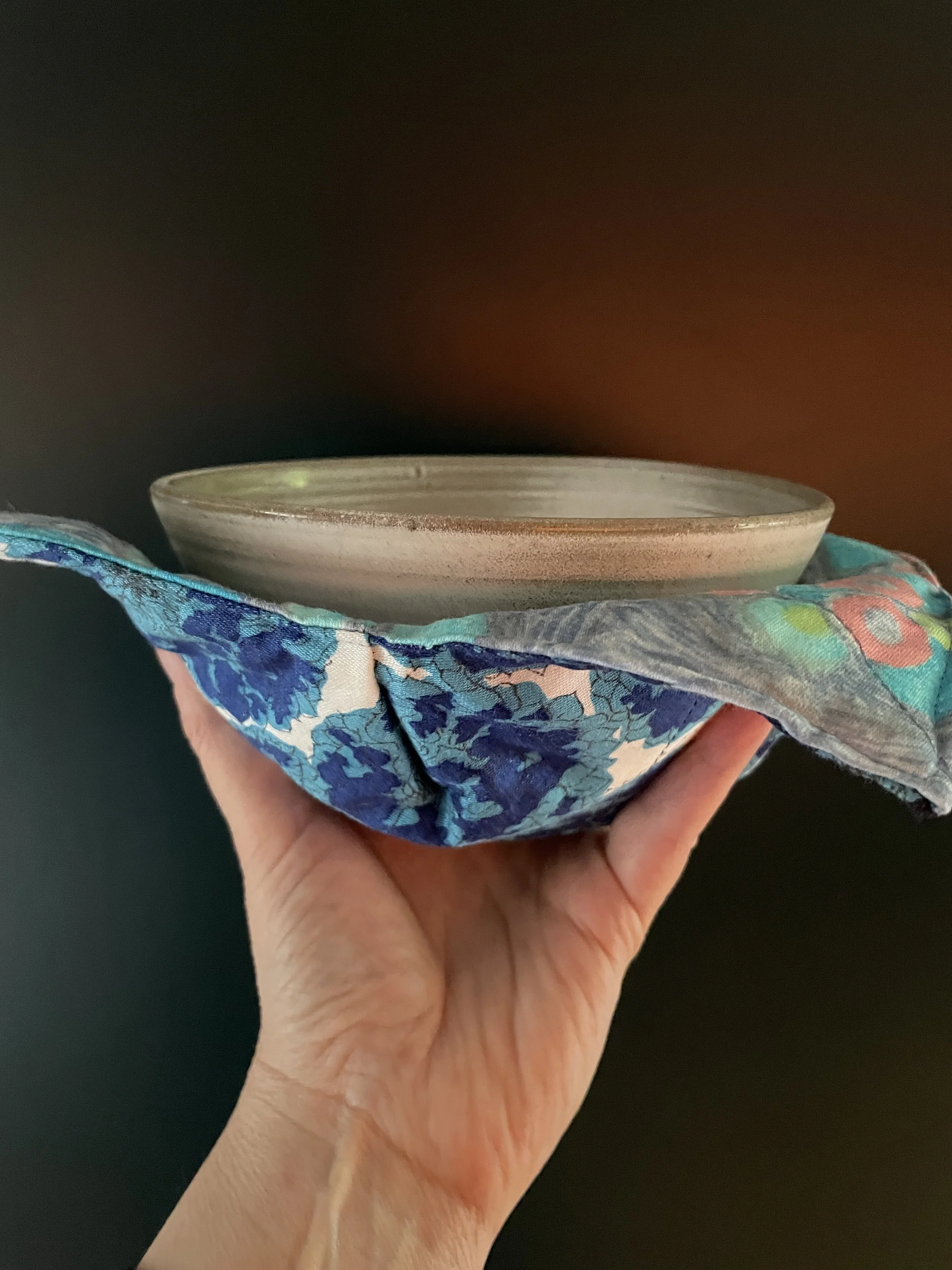 A hand holding a ceramic bowl on top of a colorful fabric bowl holder against a dark background.