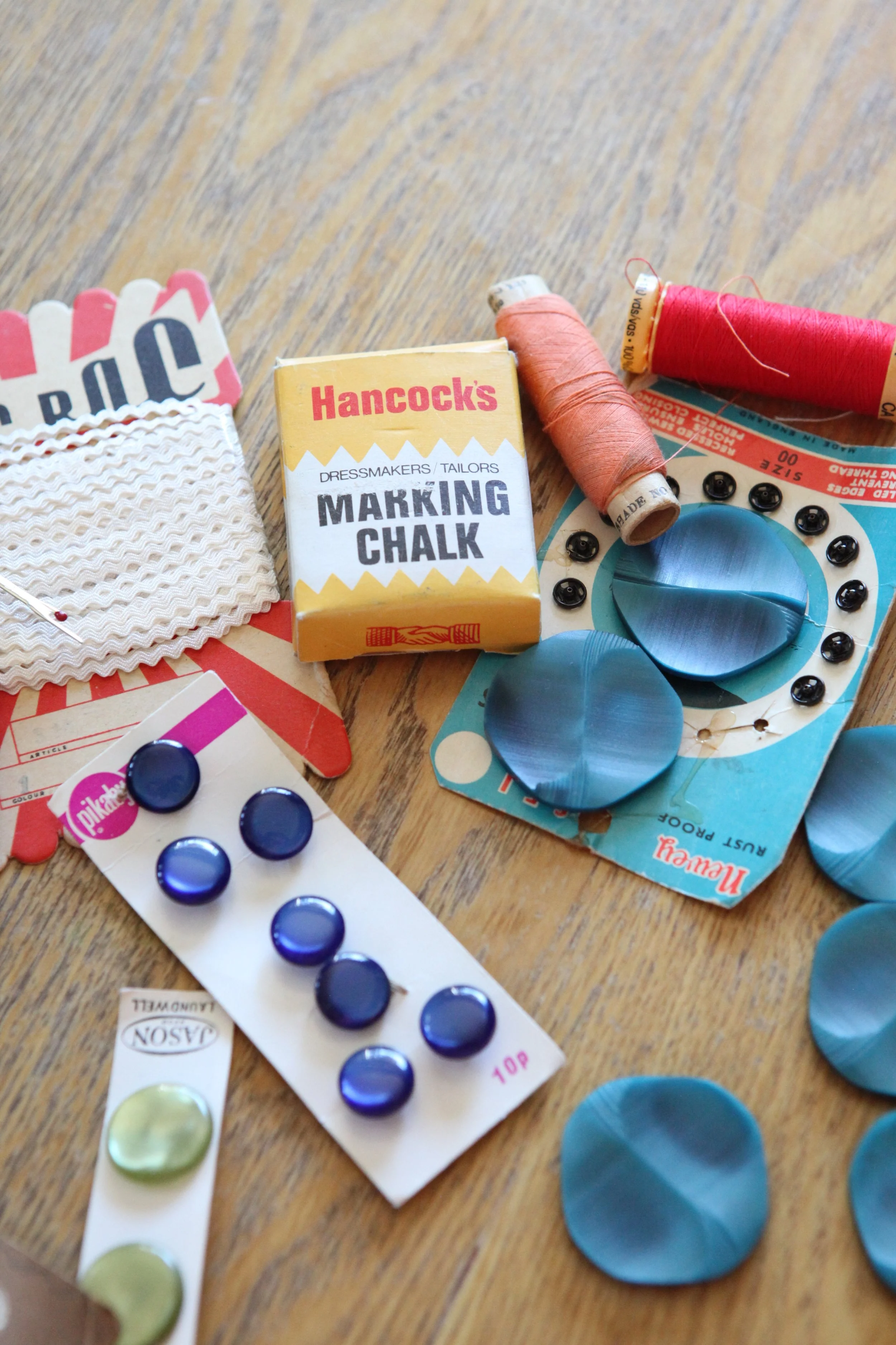 Assorted sewing and craft supplies on a wooden surface, including spools of thread, ric rac trim on a card board, buttons, marking chalk and poppers.