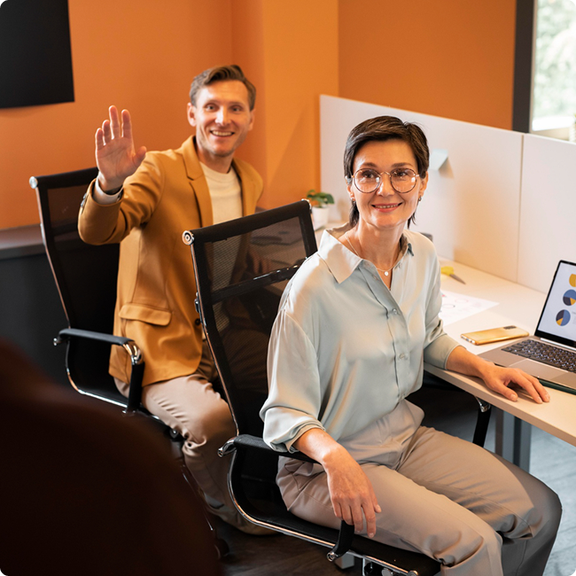 Two smiling colleagues, a man and a woman, sitting in an office, with the man waving at the camera, surrounded by office supplies and a laptop with charts on the screen.