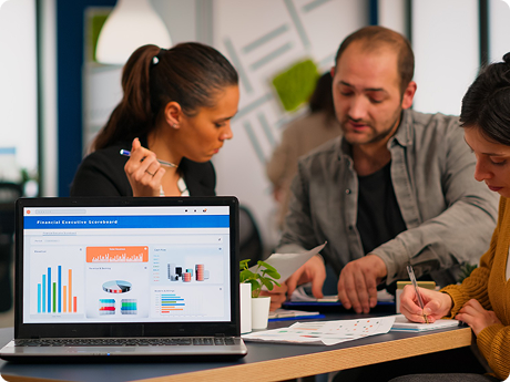 Group of three people collaborating on a business project, with a laptop displaying financial charts and graphs on a table in an office.