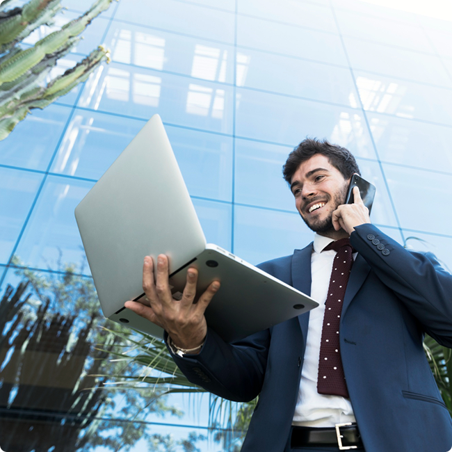 Businessman in a suit holding a laptop and talking on the phone outside a modern glass building.