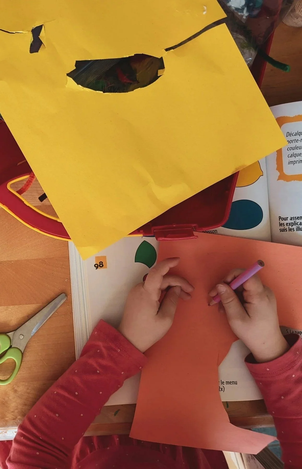 A child wearing a red long-sleeve shirt is coloring with a pink marker on orange paper. A pair of green-handled scissors and a yellow paper with a black hole cut-out are on the table. An open activity book is partially visible nearby.