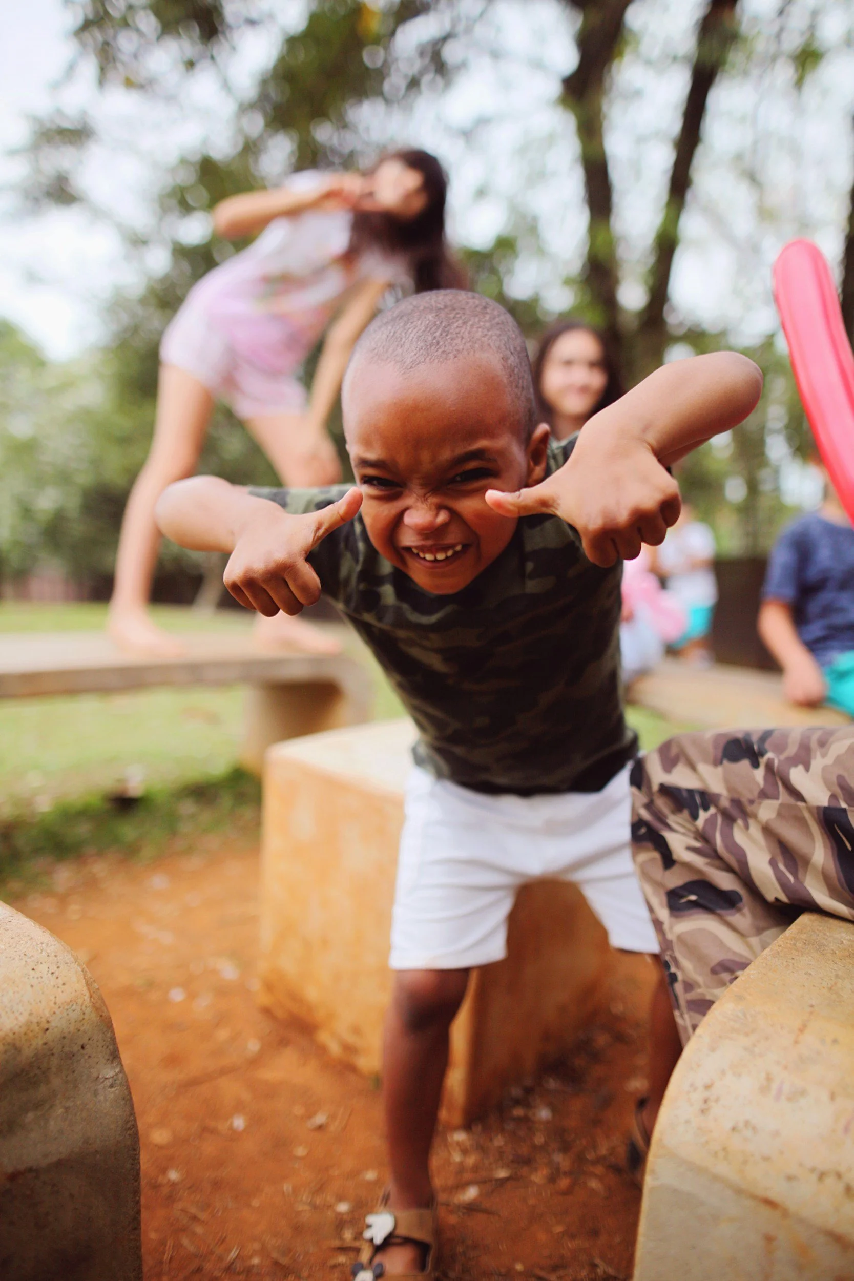 A young boy with a buzz cut, smiling and pointing at himself, leaning forward with his thumbs up. Behind him are two women, one in a pink dress and one in a dark shirt, all outdoors in a park with trees and playground equipment.