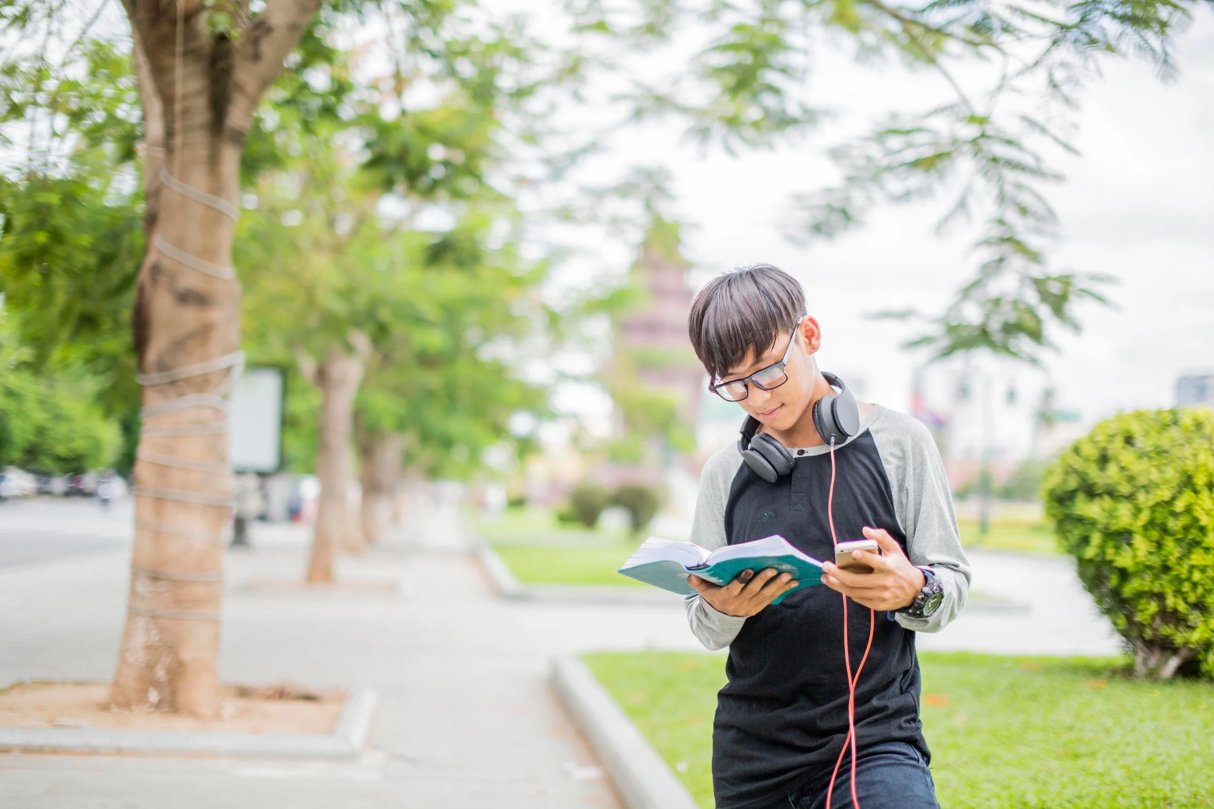 Young man with glasses, headphones, and a watch walking in a park, looking at a book and a smartphone.