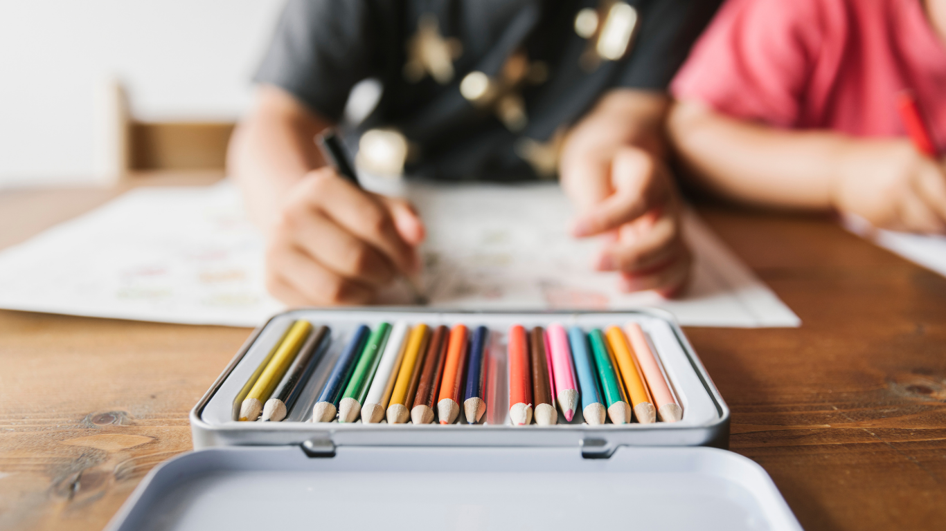 Colorful markers inside an open tin box on a wooden table with two children drawing in the background.