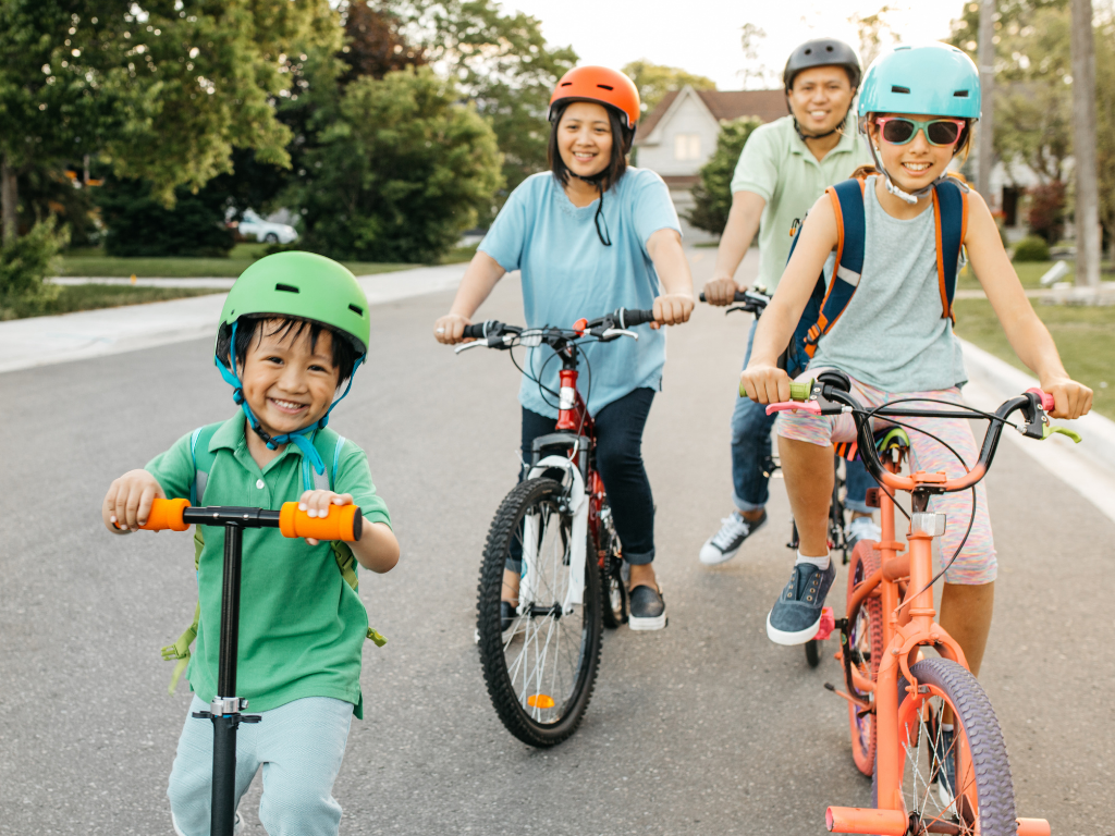 A group of four children and one adult riding bikes and scooters on a suburban street, all wearing helmets and smiling.