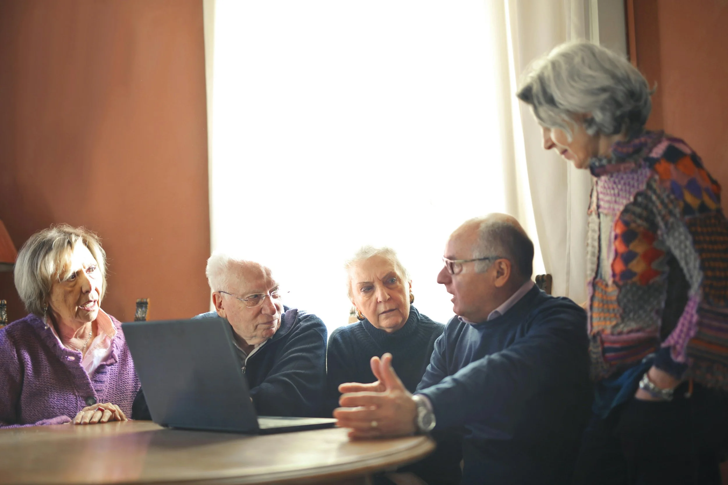 A group of five seniors engaged in a serious discussion around a table, with a laptop open in front of them, in a well-lit room with brown walls and a large window.