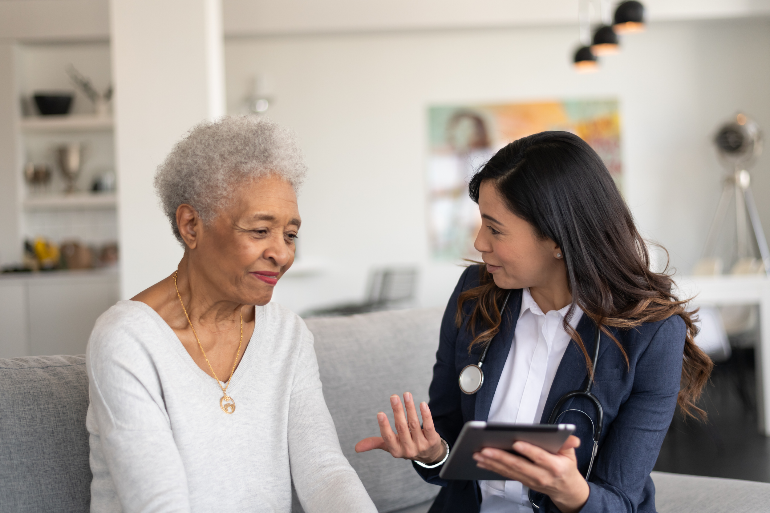 A young female doctor consulting with an elderly woman in a modern, well-lit living room. The doctor is holding a tablet and gesturing while the elderly woman listens attentively.