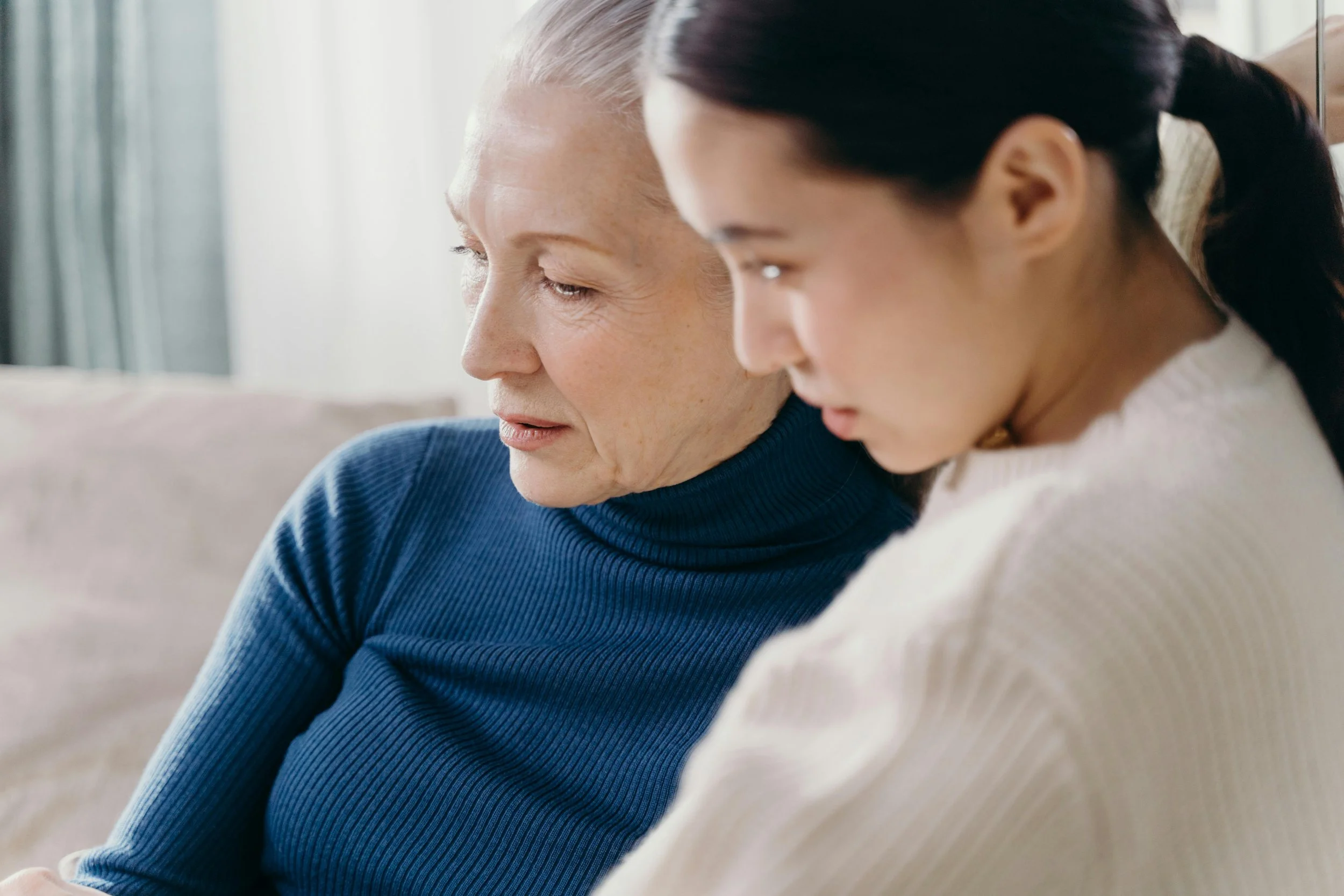 Two women, one older with gray hair in a blue turtleneck, and one younger with dark hair in a white sweater, sitting closely and looking down together in a cozy indoor setting.