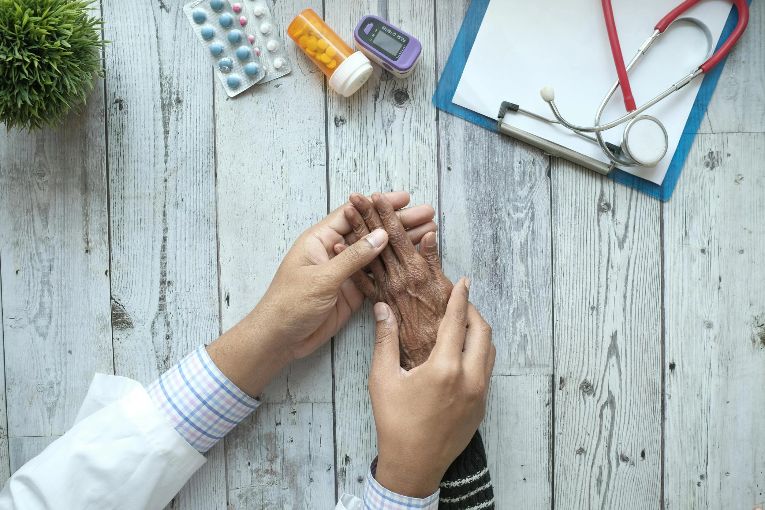 A healthcare professional holding an elderly person's hand on a doctor’s desk with prescription pills, a stethoscope, and a blood glucose meter on a wooden surface.