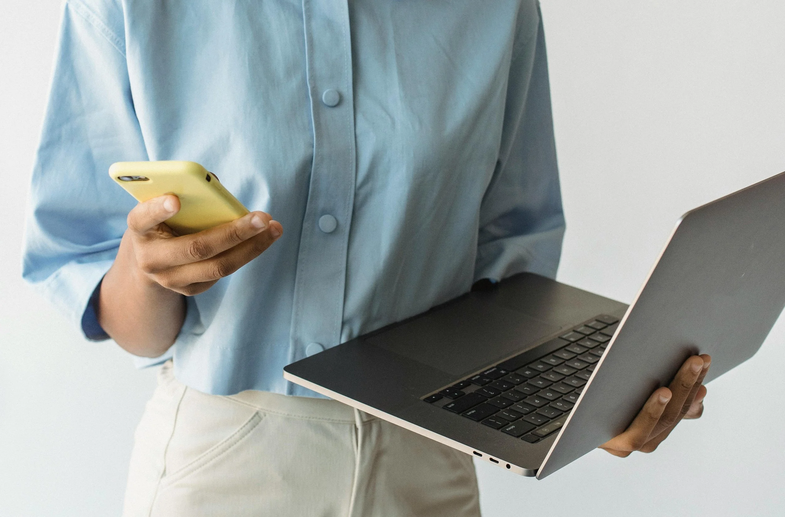 Person holding a yellow smartphone in one hand and a silver laptop in the other, wearing a light blue button-up shirt and light-colored pants.