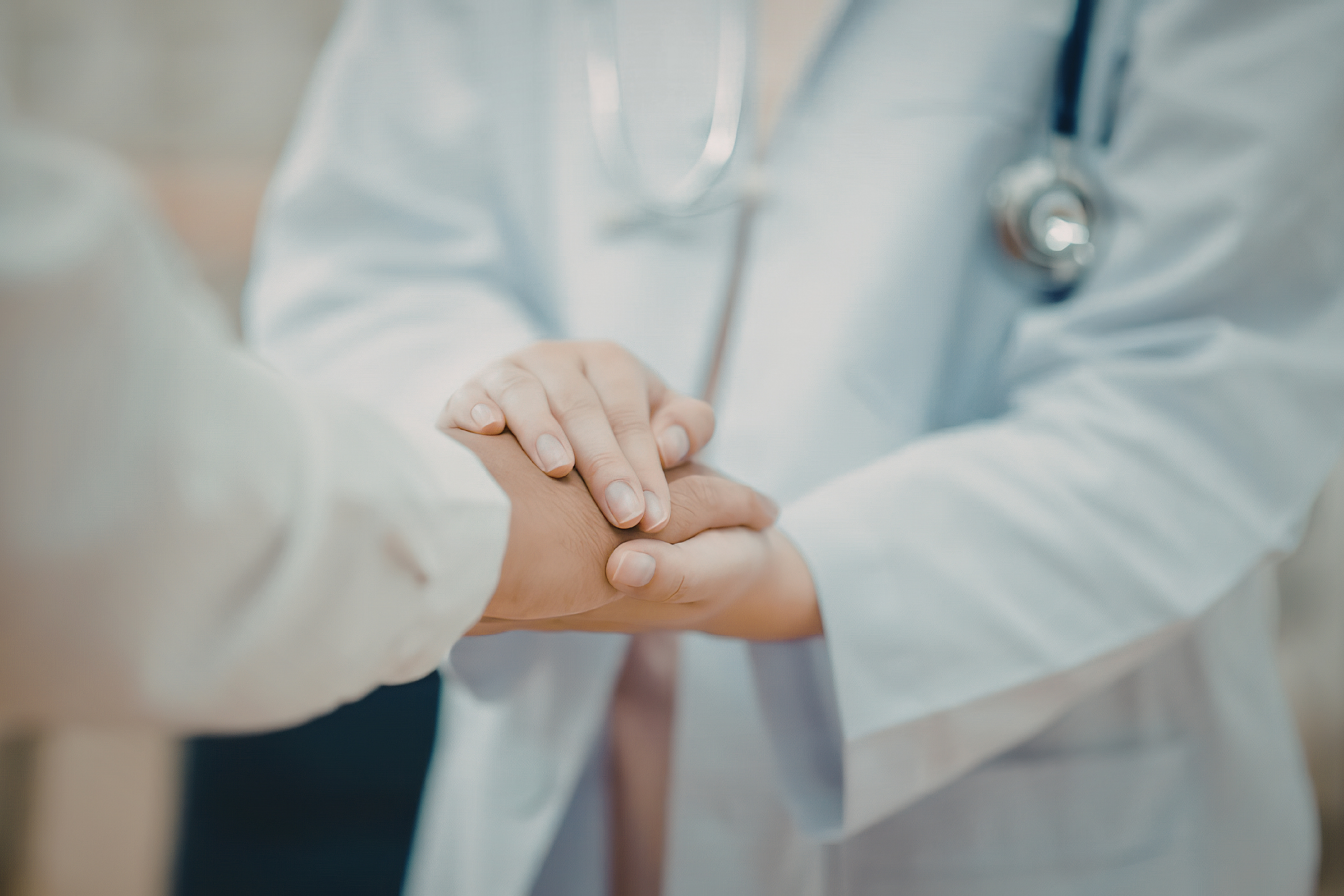 A healthcare professional wearing a white coat is holding the hand of a patient, with a stethoscope around their neck.